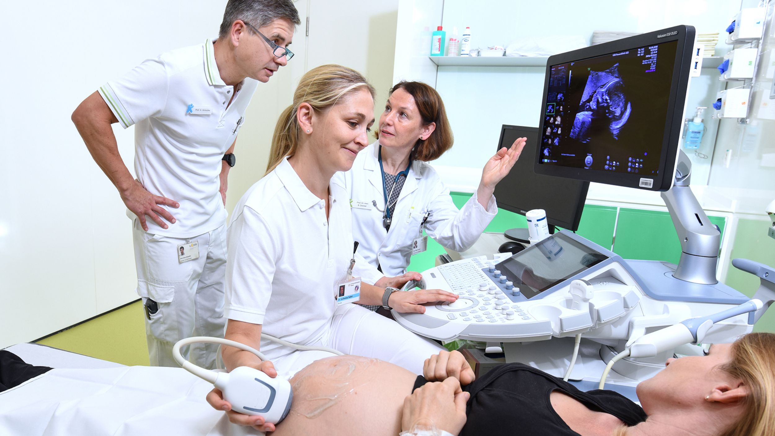 Eine schwangere Patientin erhält einen Ultraschall von einer Ärztin. Im Hintergrund sehen sich ein Arzt und Ärztin von der Neonatologie das Bild an.
