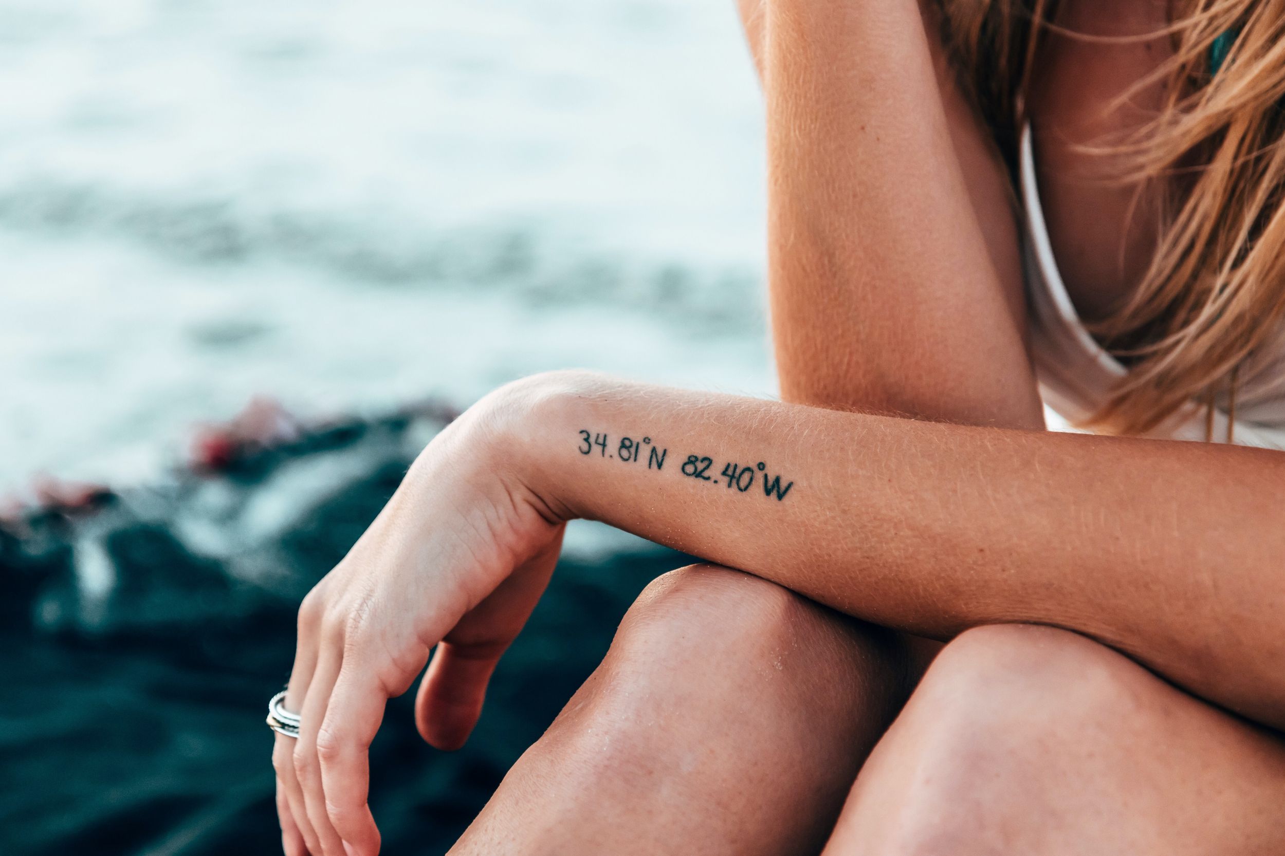Frau mit kleinem Tattoo am Arm sitzt am Strand im Hintergrund sieht man das Meer