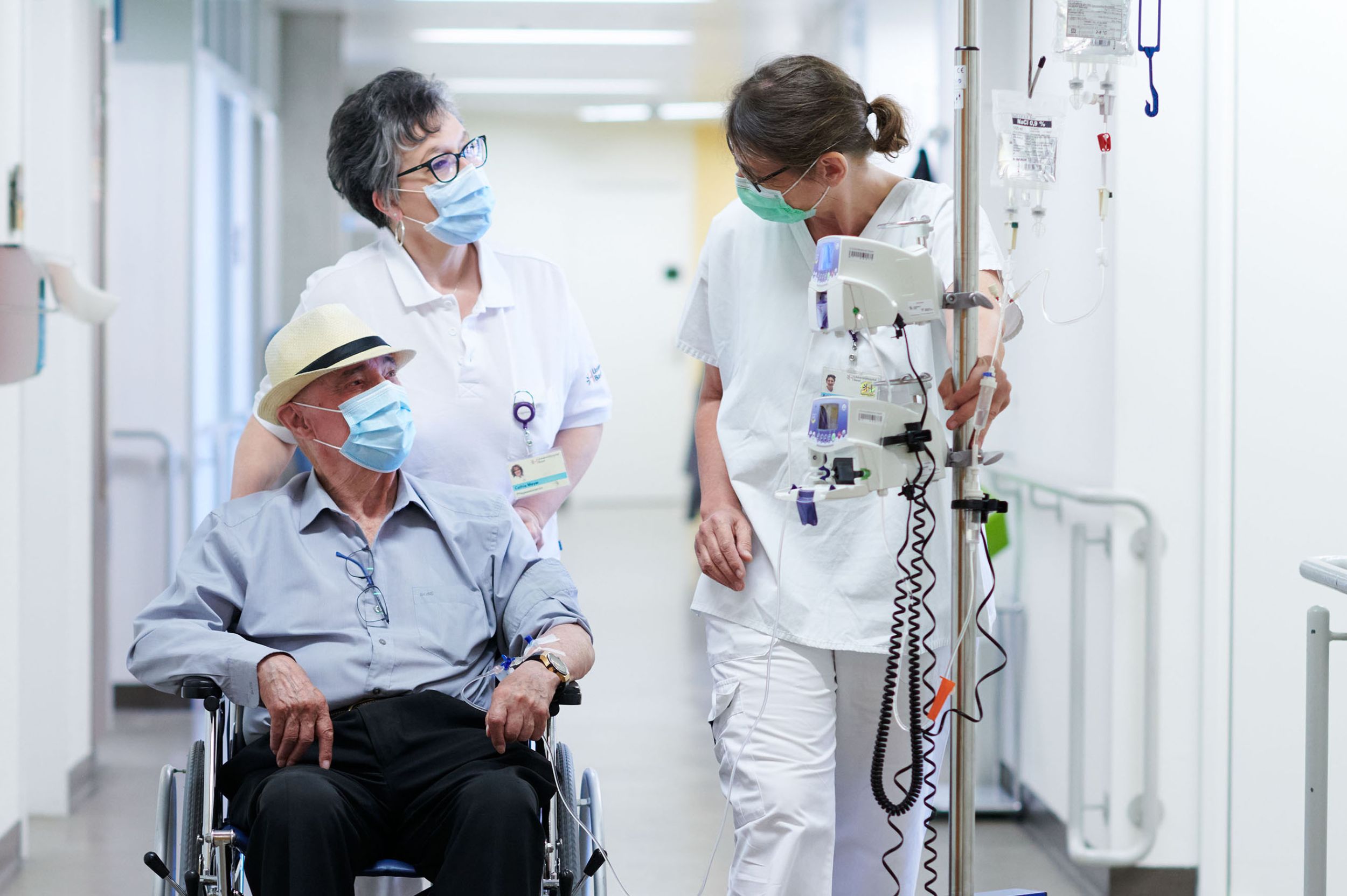 Pflegefachperson begleitet einen Patienten im Rollstuhl gemeinsam mit einer Angehörigen durch den Gang
