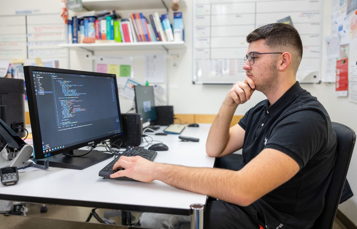 Informatiker EFZ, Fachrichtung Applikationsentwicklung, in Ausbildung sitzt mit schwarzem Polo-Shirt sitzt am Computer und programmiert