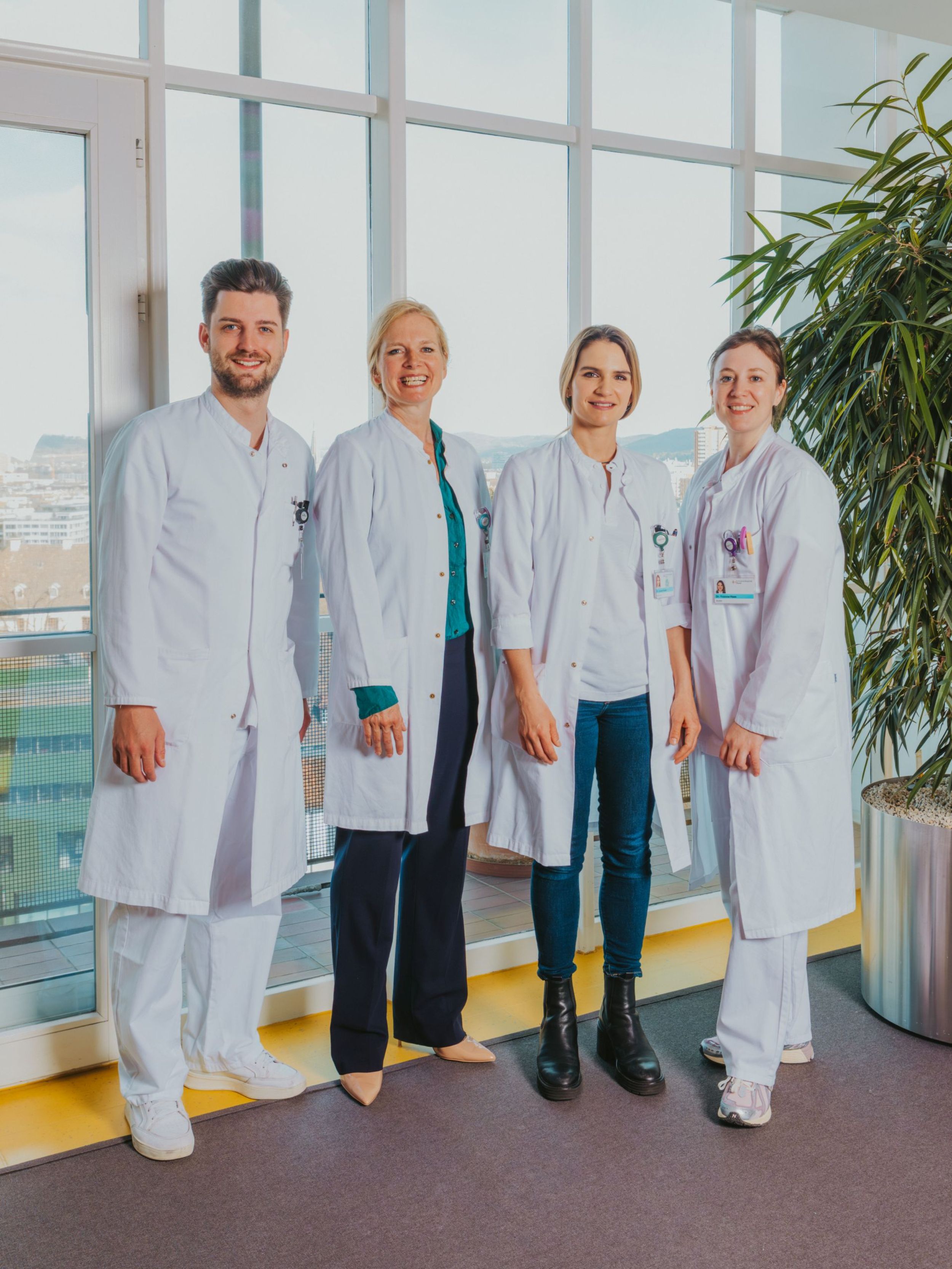 Vier medizinische Fachpersonen in weissen Kitteln stehen nebeneinander vor grossen Fenstern und lächeln in die Kamera; im Hintergrund ist eine Stadtansicht zu sehen.