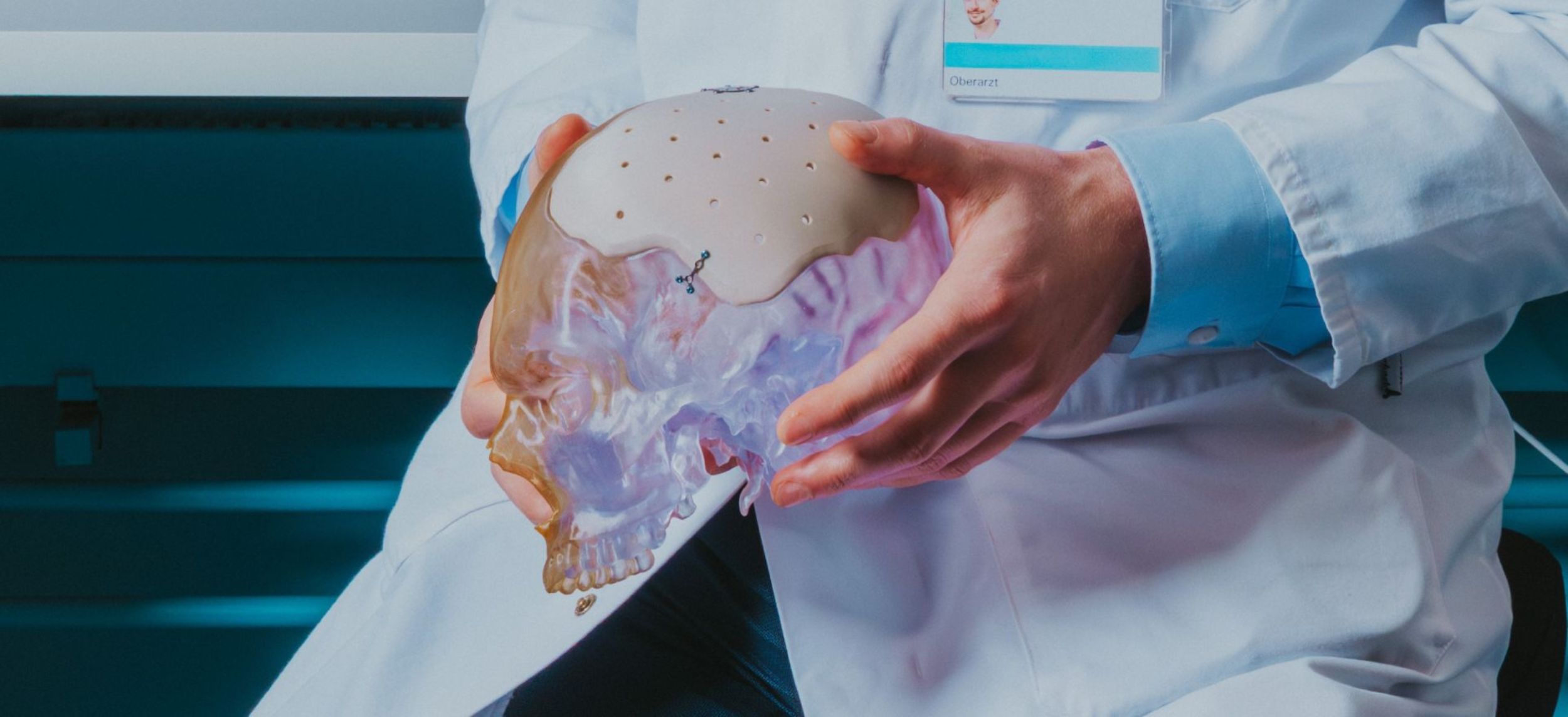 Ein Arzt im weissen Kittel hält ein anatomisches Schädelmodell in den Händen und zeigt Details am Kopf.