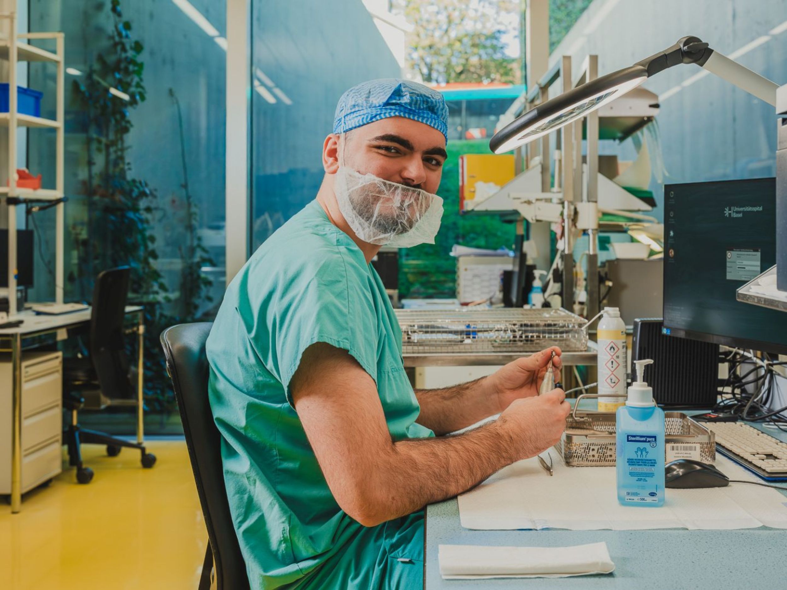 Medizinischer Mitarbeiter in Berufskleidung sitzt an einem Arbeitsplatz und bereitet Instrumente auf, während er zur Kamera blickt; auf dem Tisch stehen Desinfektionsmittel und technische Geräte.