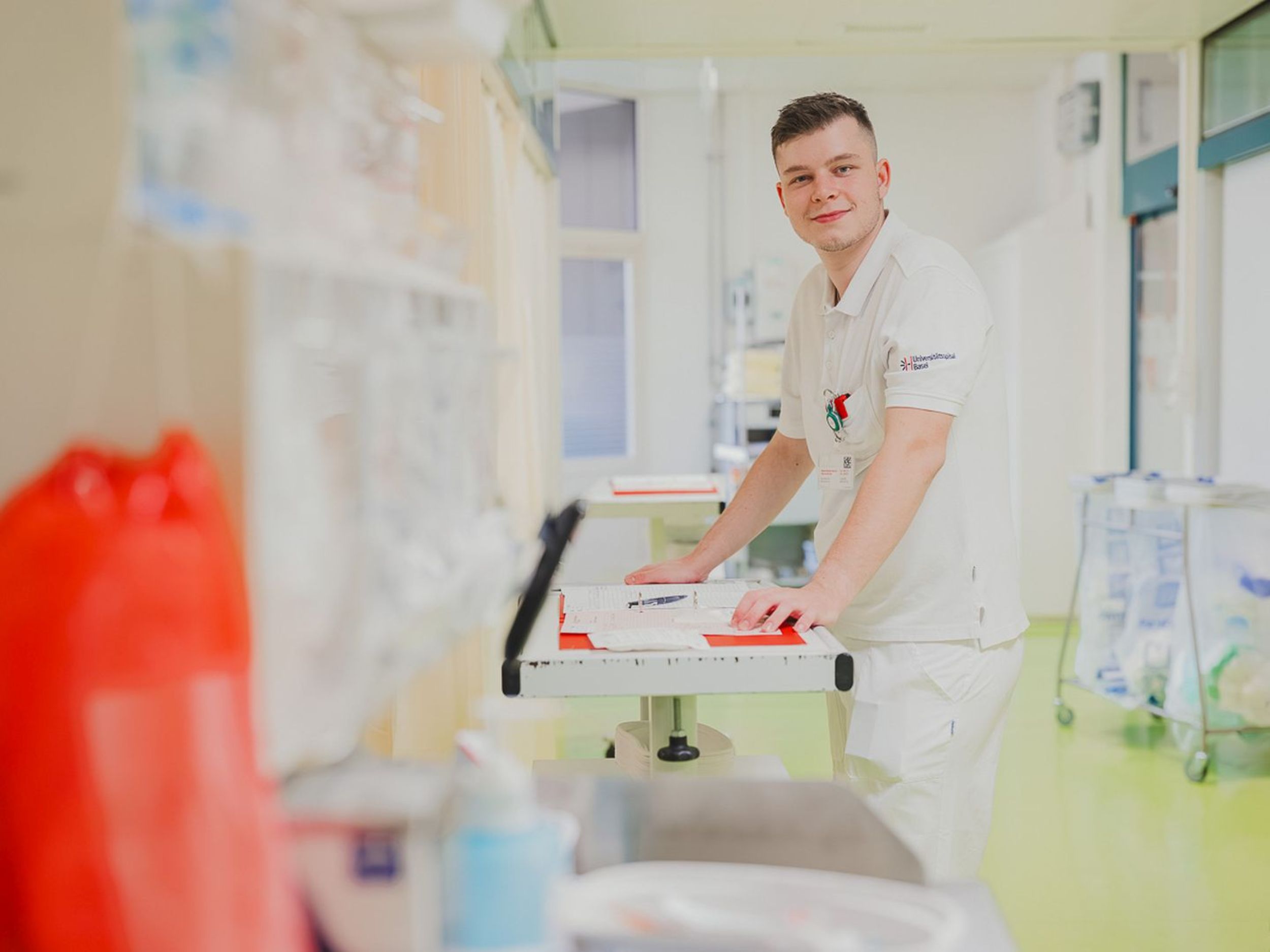 Une scène dans un environnement hospitalier montre du personnel médical en tenue professionnelle blanche s'occupant d'un patient sur un lit. Un homme pousse le lit à travers un couloir, tandis qu'une autre personne portant un masque accompagne le lit. Au premier plan, un autre membre du personnel est assis à une table. L'environnement est clairement structuré, avec des appareils médicaux, des moniteurs et des systèmes de rangement. La scène montre un service de soins intensifs ou d'urgence.