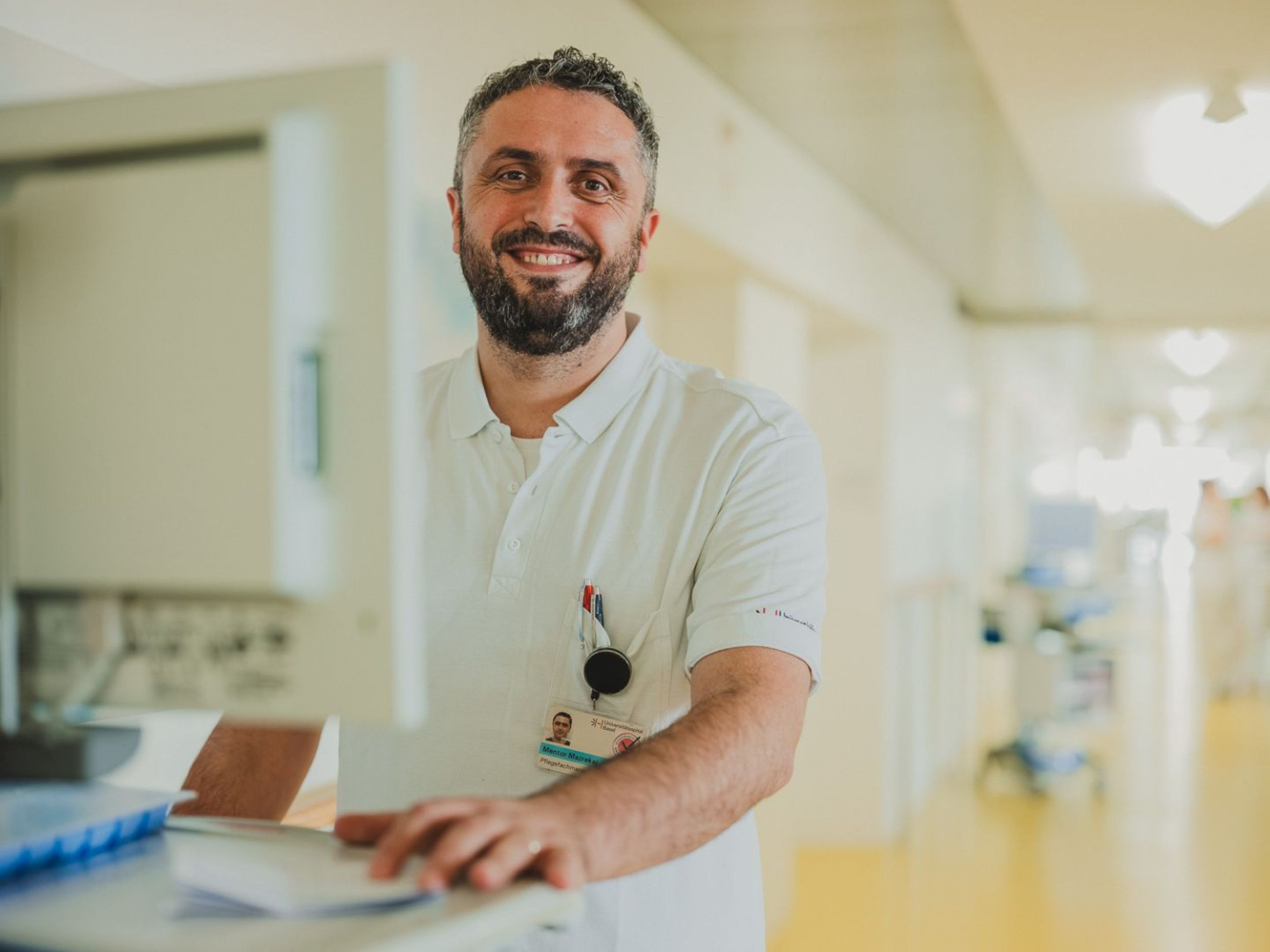 Une femme en tenue professionnelle blanche se penche avec concentration sur une patiente dans un environnement médical. L'arrière-plan montre des étagères murales avec des gants bleus et d'autres ustensiles médicaux. La scène montre une situation de soins ou de traitement dans un établissement médical professionnel.