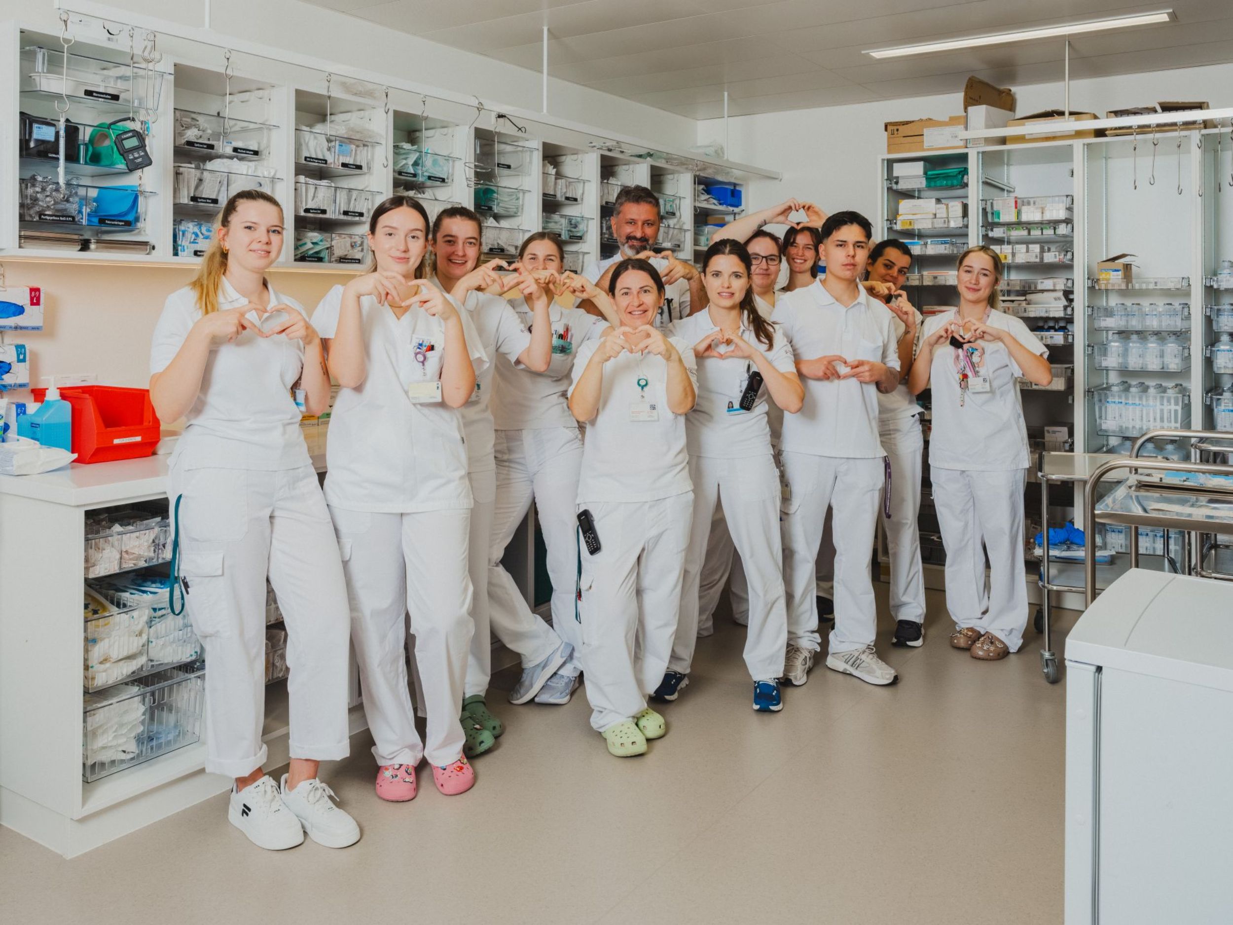 Ein Team von Pflegefachpersonen steht gemeinsam in einem Materialraum und formt mit den Händen Herzsymbole in Richtung Kamera.