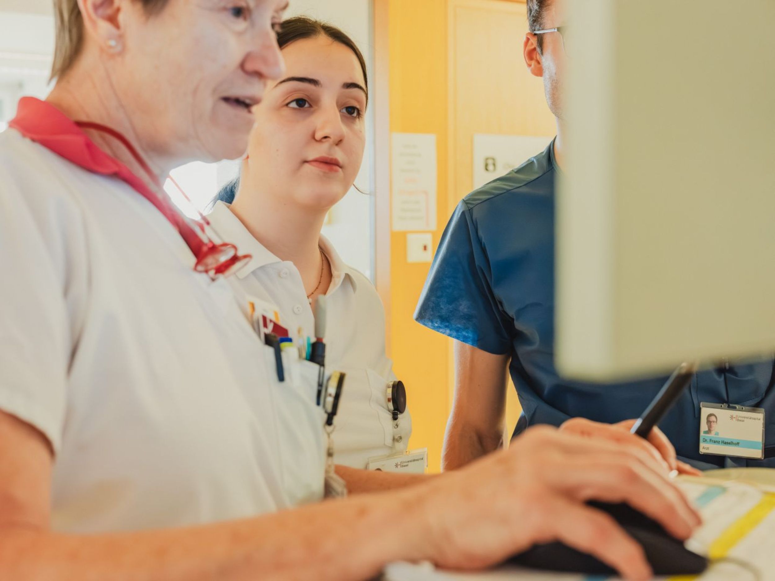 Zwei Pflegefachpersonen und ein Arzt stehen gemeinsam vor einem Computer und besprechen etwas am Bildschirm; eine Person bedient die Maus, während die anderen aufmerksam zuschauen.