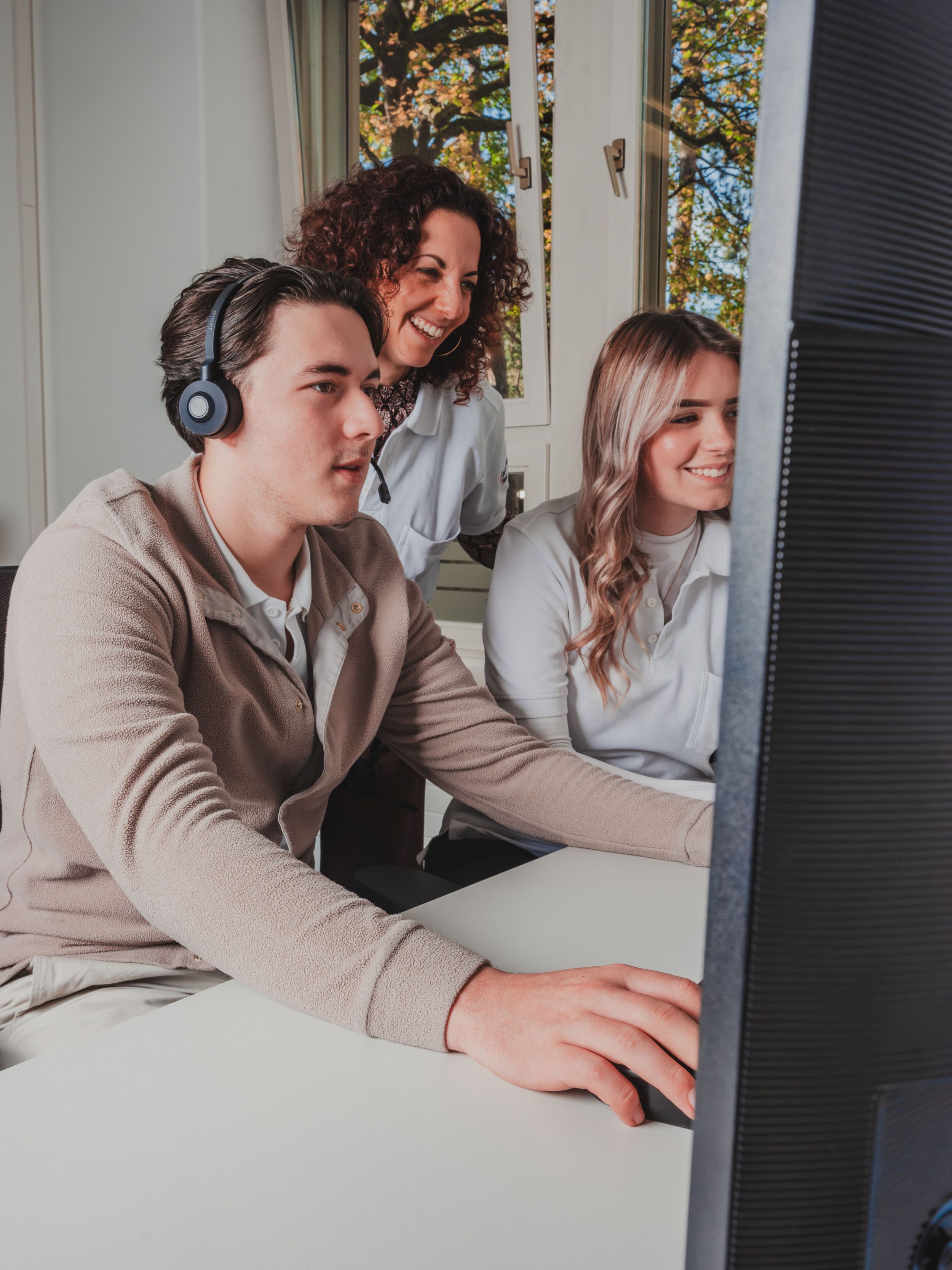 Drei junge Personen arbeiten gemeinsam am Computer: Eine Person mit Headset sitzt am Bildschirm und bedient die Maus, während zwei Kolleginnen danebenstehen, lächeln und unterstützend mitarbeiten; im Hintergrund fällt Tageslicht durch ein Fenster.