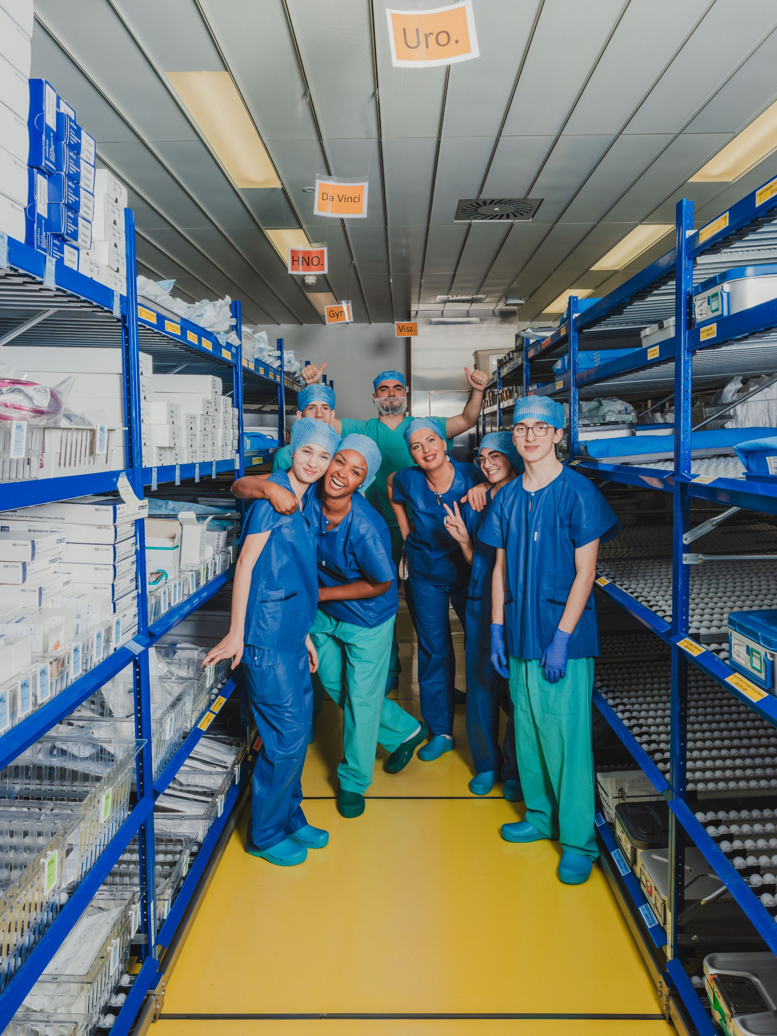 Gruppe von medizinischen Mitarbeitenden in OP-Kleidung steht lächelnd zwischen Regalen mit steril verpackten Materialien in einem Lagerraum; über ihnen hängen Beschriftungen für verschiedene Fachbereiche.