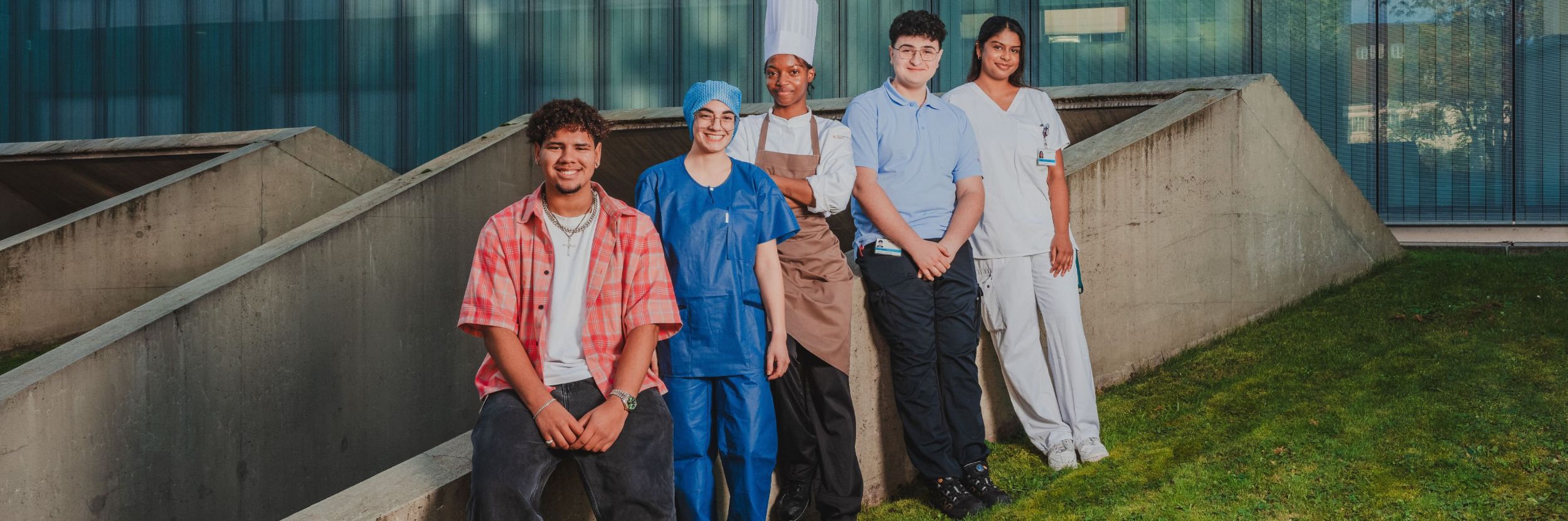 Fünf junge Menschen in unterschiedlichen Berufsbekleidungen stehen und sitzen gemeinsam im Freien vor einem modernen Gebäude mit Glasfassade und schauen in die Kamera.