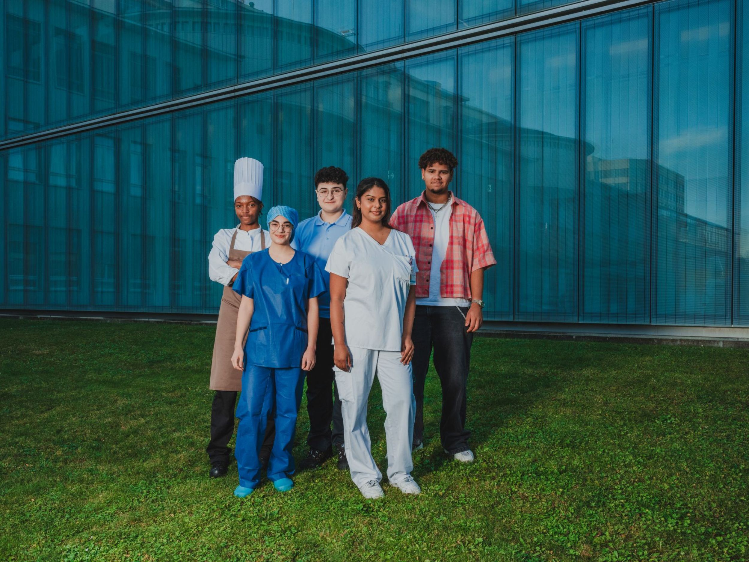Fünf junge Menschen in unterschiedlichen Berufsbekleidungen (u. a. Pflege, Gastronomie und Alltag) stehen gemeinsam vor einem modernen Gebäude mit Glasfassade auf einer Grünfläche und schauen in die Kamera.