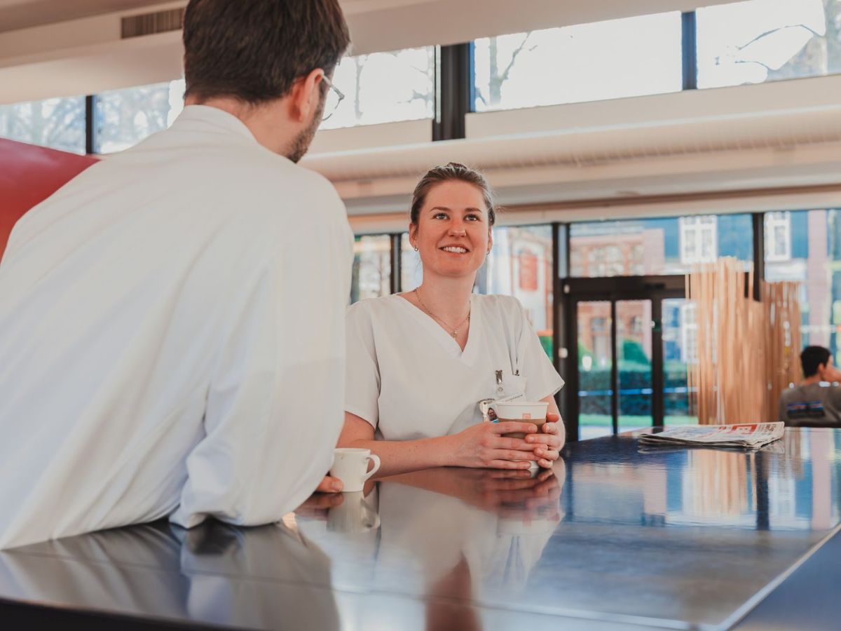 Zwei Mitarbeitende im Gesundheitswesen stehen an einem Tisch, trinken Kaffee und unterhalten sich in entspannter Atmosphäre.