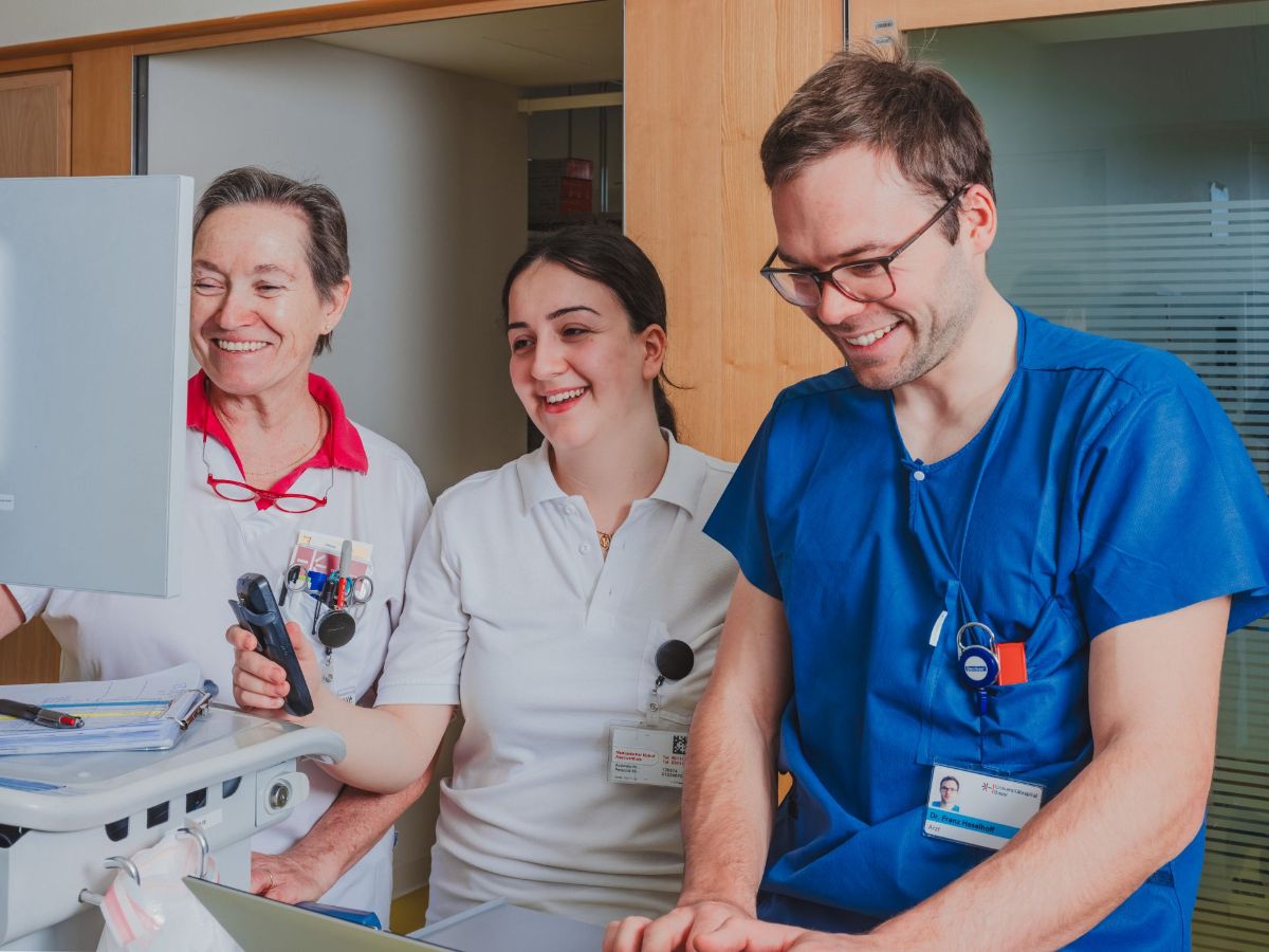 Drei Mitarbeitende im Gesundheitswesen stehen zusammen an einem mobilen Arbeitsplatz, schauen auf einen Bildschirm und lachen gemeinsam.