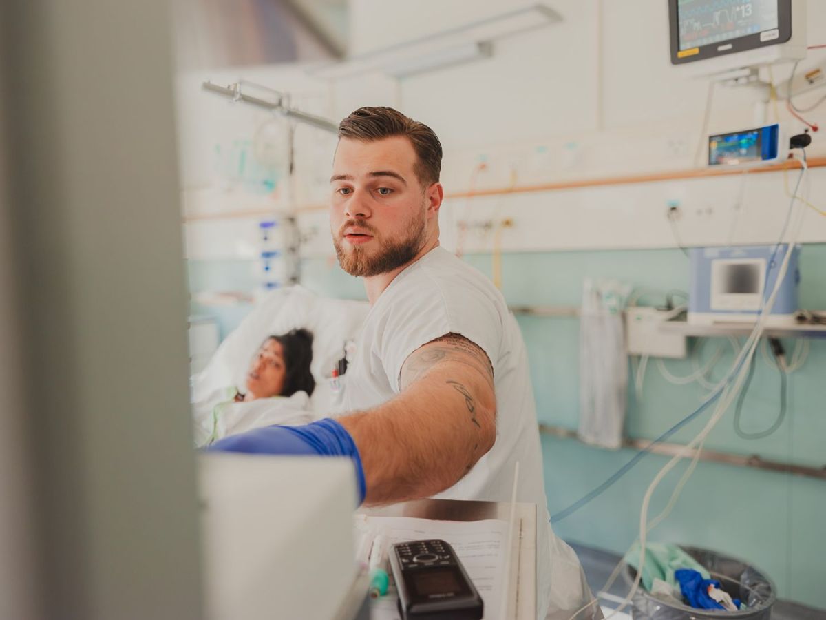 Un homme aux cheveux attachés en arrière et portant une boucle d'oreille, vêtu d'une chemise blanche, se tient à une table et écrit avec concentration sur du papier à l'aide d'un stylo. Dans la poche de sa chemise sont rangés des stylos et des documents. L'arrière-plan montre un environnement de travail lumineux, légèrement flou, avec des ustensiles médicaux.