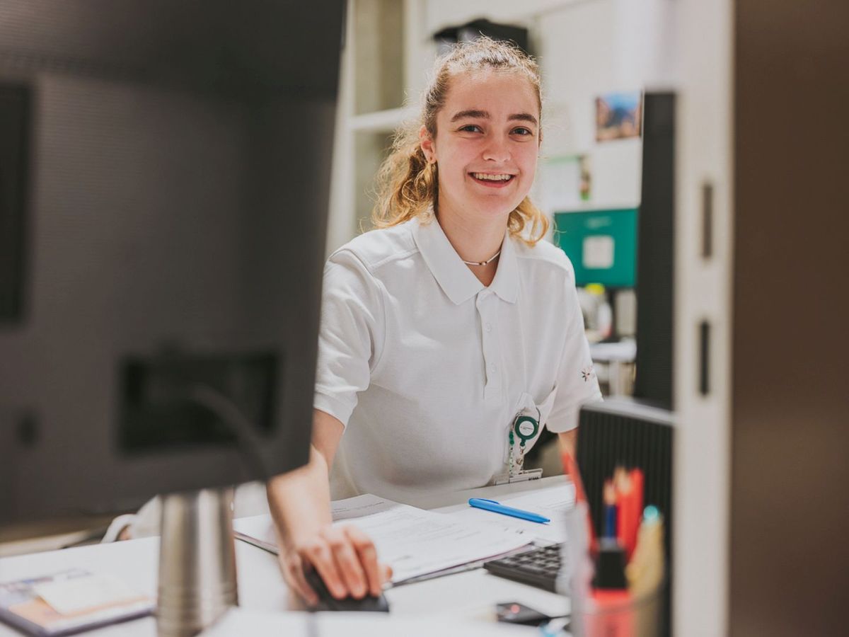 Une femme vêtue d'une tenue professionnelle blanche se tient dans une salle médicale et utilise un appareil électronique avec plusieurs câbles et écrans. Elle semble concentrée lorsqu'elle appuie sur une touche. En arrière-plan, on aperçoit une armoire ainsi qu'un tableau abstrait coloré sur le mur. L'environnement montre un poste de travail médical professionnel.