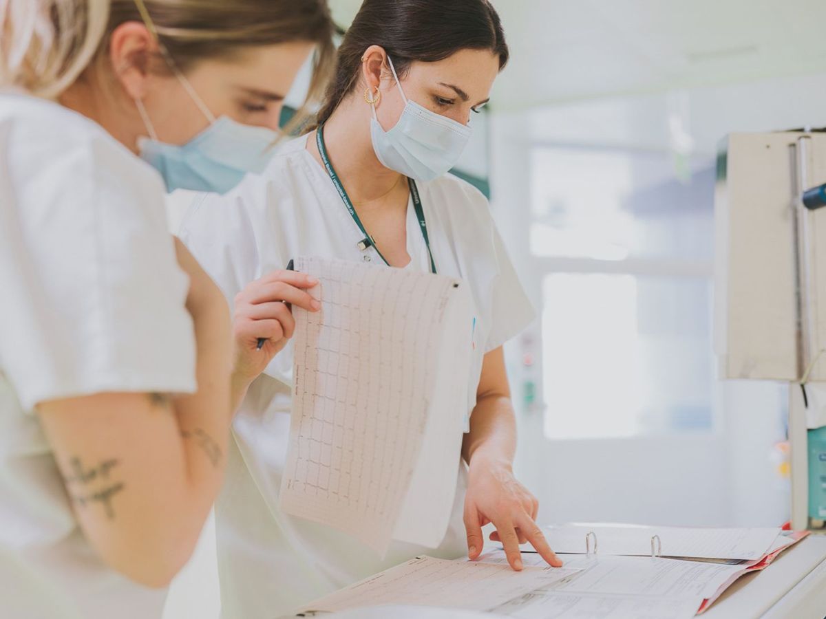 n équipe de trois femmes en vêtements professionnels blancs et masques médicaux se tient devant un panneau mural portant des inscriptions telles que 'gestion de la journée'. Une femme, debout au milieu, porte des lunettes et un badge et semble écouter attentivement. Une autre montre le tableau d'une main, tandis que la troisième femme lui tourne le dos. La scène montre une réunion concentrée dans un environnement de travail professionnel.