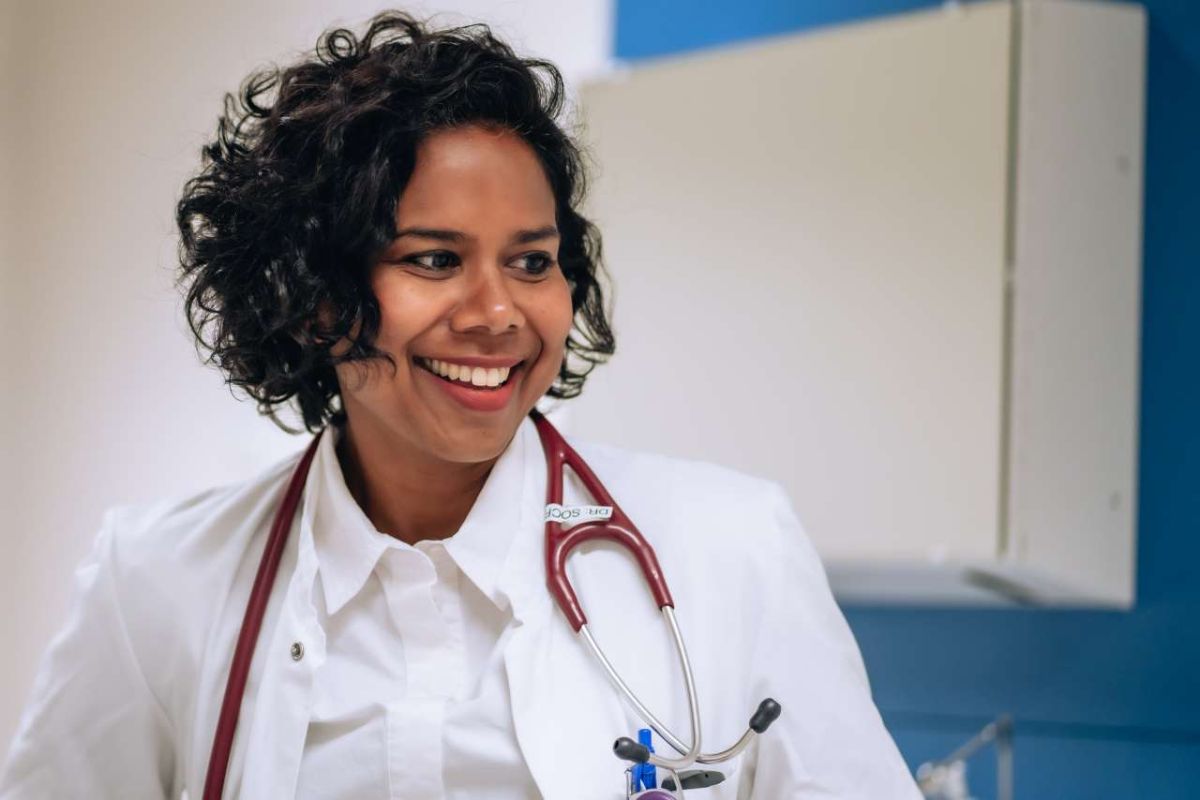 Ärztin in weißem Kittel mit Stethoskop um den Hals, lächelnd in einem Behandlungsraum