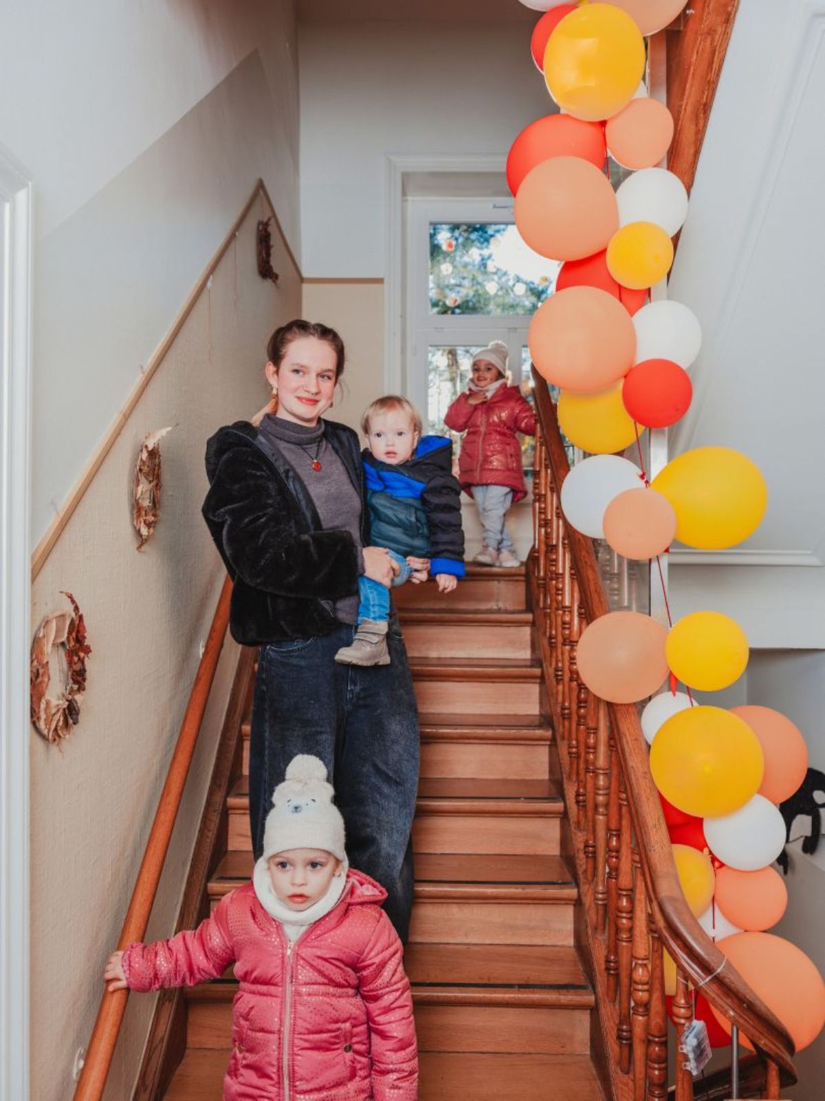 Eine Betreuerin steht mit einem Kleinkind auf einer Treppe, während weitere Kinder die Treppe hinuntergehen; daneben sind bunte Ballons befestigt.