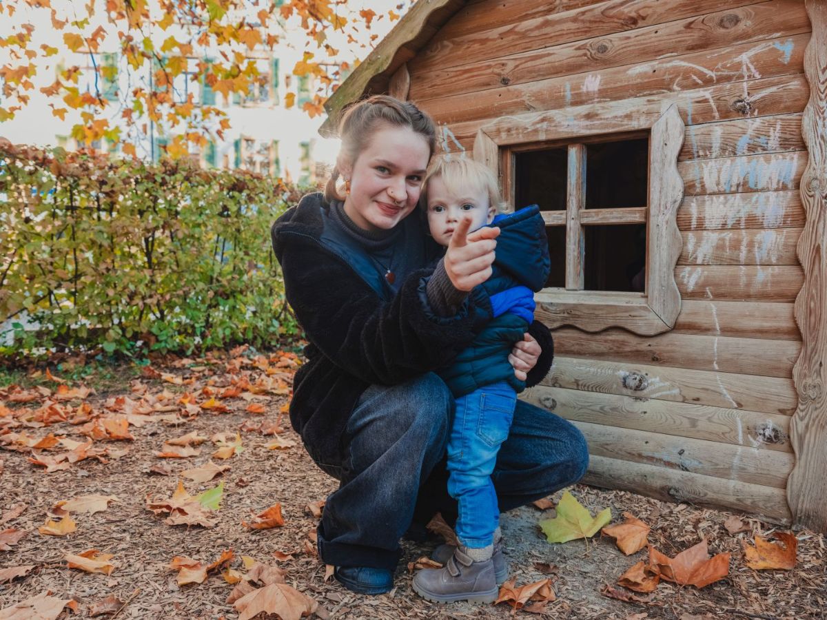 Eine junge Frau hockt neben einem Kleinkind vor einem kleinen Holzspielhaus im Freien und zeigt mit dem Finger auf etwas.