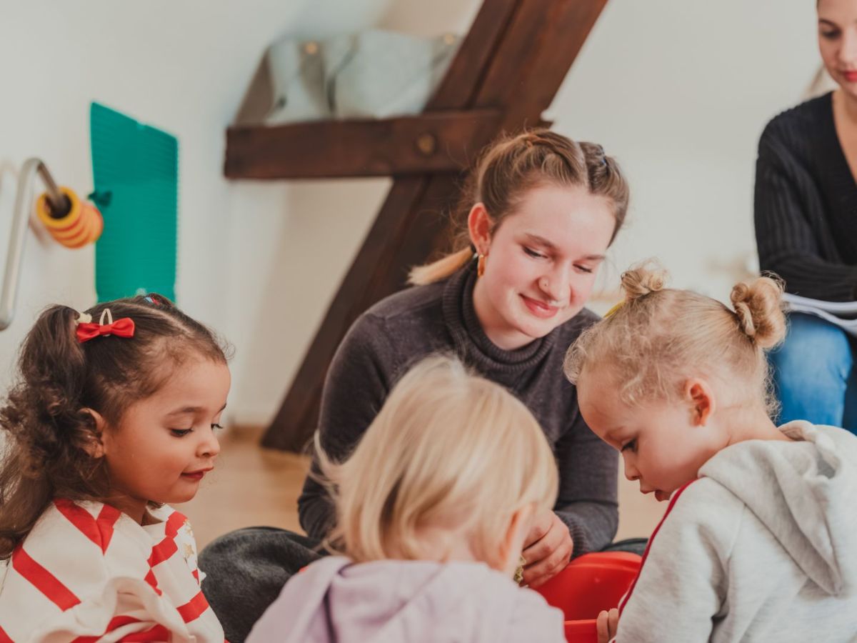 Eine Betreuerin sitzt mit mehreren Kleinkindern auf dem Boden und beschäftigt sich gemeinsam mit ihnen beim Spielen.