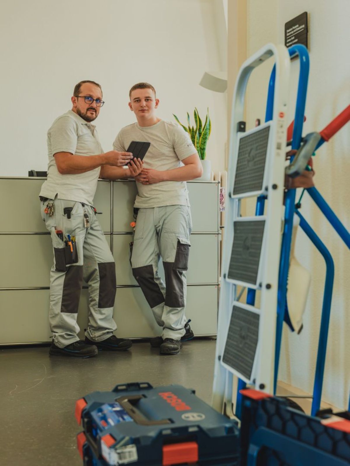 Zwei Mitarbeitende im Bereich Betriebsunterhalt stehen in einem Innenraum, halten ein Tablet in der Hand und schauen in die Kamera; im Vordergrund stehen Werkzeuge und eine Leiter.