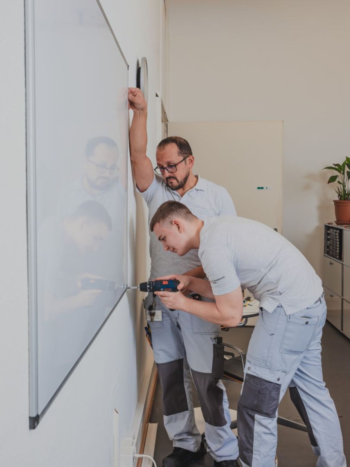 Zwei Mitarbeitende im Betriebsunterhalt montieren gemeinsam etwas an einer Wand; eine Person arbeitet mit einer Bohrmaschine, die andere schaut zu.