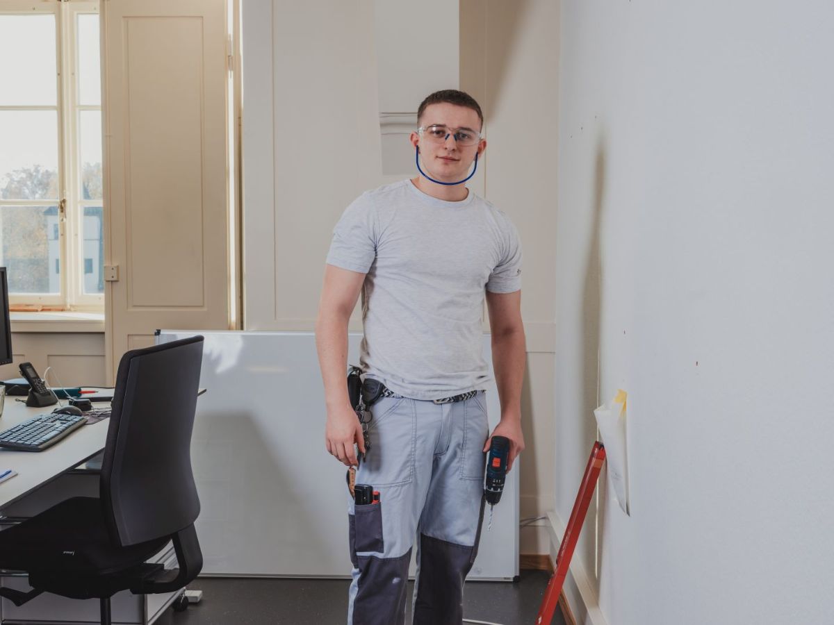 Ein Mitarbeitender im Betriebsunterhalt steht in einem Innenraum mit Schutzbrille und Bohrmaschine vor einer Wand.