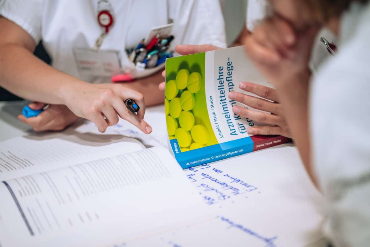 Pflegefachfrauen in Ausbildung lernen gemeinsam mit Fachliteratur, zeigen auf Textstellen und halten ein Lehrbuch zur Arzneimittellehre.