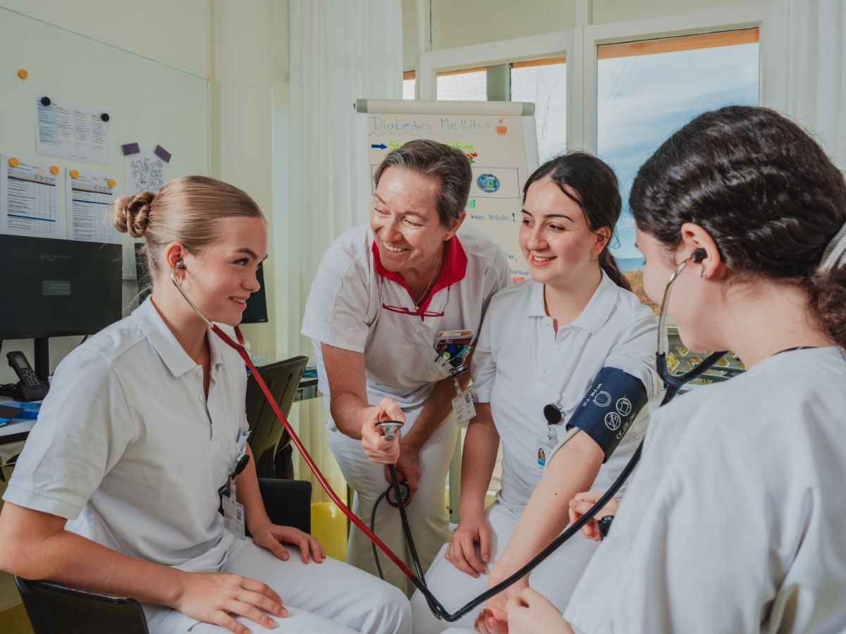 Une jeune femme en tenue professionnelle blanche se tient debout, souriante, dans une pièce lumineuse. Elle porte un badge avec l'inscription 'Hôpital universitaire de Bâle' sur la manche de sa chemise. En arrière-plan, on aperçoit une fenêtre, une commode à tiroirs vert clair et des ustensiles médicaux tels que des désinfectants. Le rayonnement amical et l'environnement professionnel créent une atmosphère de travail positive.