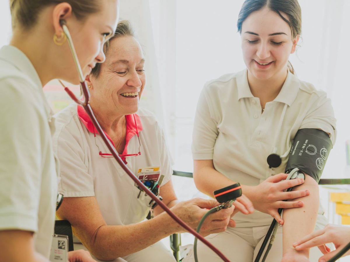 Drei Pflegefachpersonen führen gemeinsam eine Blutdruckmessung durch: Eine hält das Messgerät, eine trägt die Manschette am Arm, und eine weitere hört mit dem Stethoskop zu; alle arbeiten konzentriert in einer hellen Umgebung.