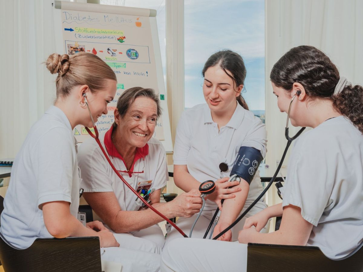 Vier Pflegefachpersonen führen gemeinsam eine Blutdruckmessung durch: Eine Person trägt die Manschette, zwei hören mit dem Stethoskop, und eine ältere Kollegin bedient das Messgerät; alle arbeiten konzentriert in einer freundlichen Lern- oder Behandlungssituation.