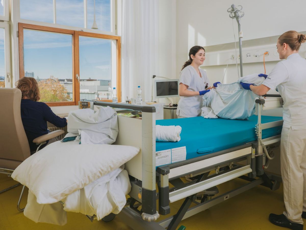 Zwei Pflegefachpersonen beziehen gemeinsam ein Spitalbett und richten es her, während im Hintergrund eine Person am Fenster sitzt; der helle Patientenzimmerbereich ist mit medizinischen Geräten und Möbeln ausgestattet.