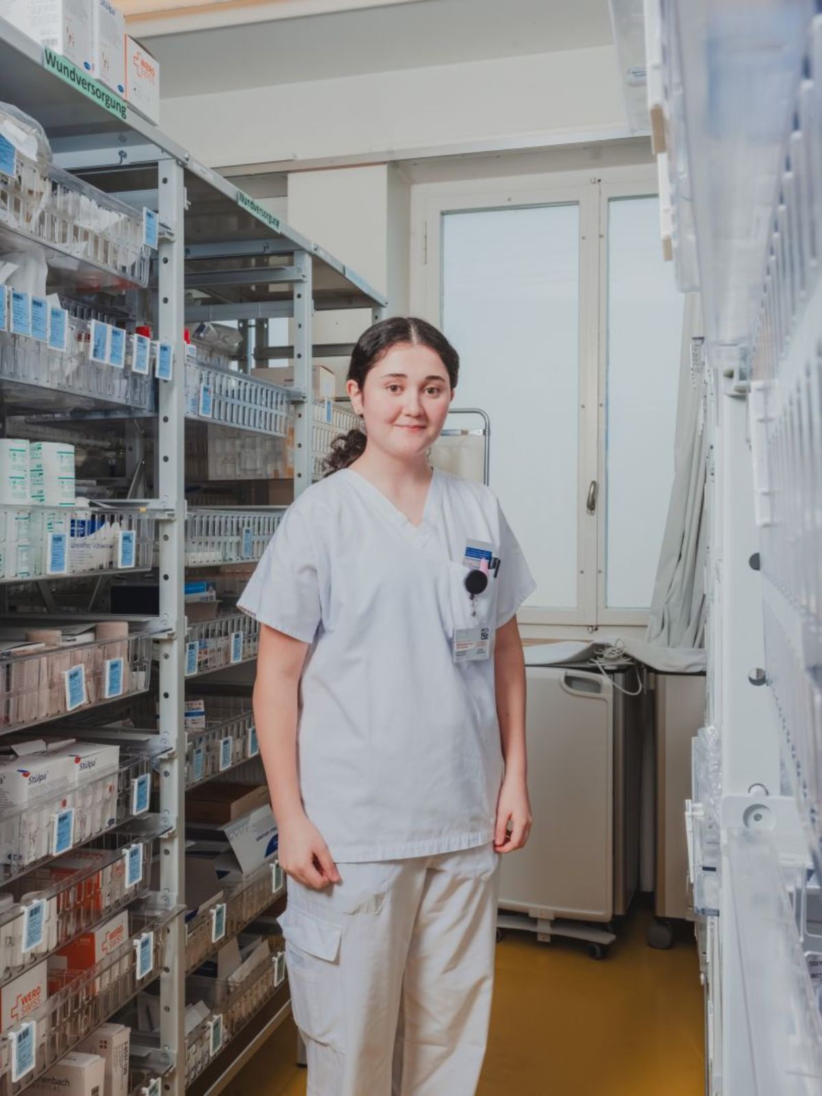 Pflegefachperson in Arbeitskleidung steht in einem Materiallager zwischen Regalen mit medizinischen Produkten und blickt in die Kamera; die Regale sind ordentlich mit beschrifteten Behältern gefüllt.