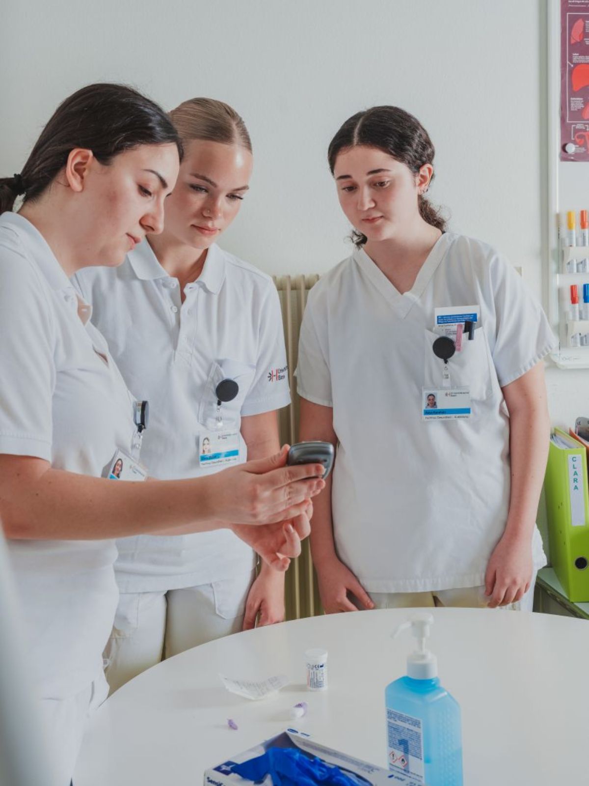 Drei medizinische Mitarbeiterinnen in Arbeitskleidung stehen gemeinsam an einem Tisch und betrachten konzentriert ein Gerät in der Hand einer Kollegin; auf dem Tisch liegen Desinfektionsmittel und medizinische Utensilien.