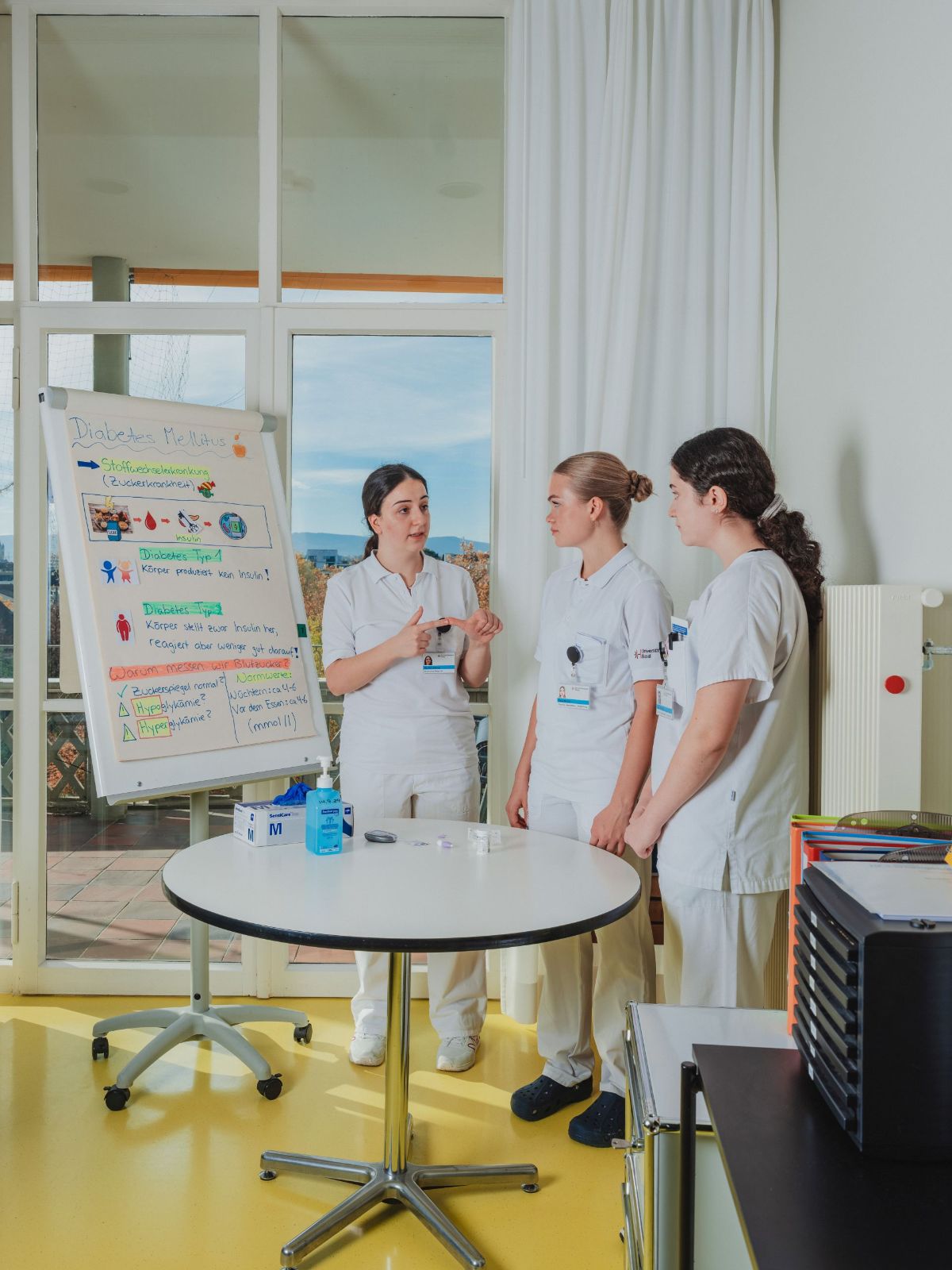 Drei Pflegefachpersonen stehen in einem hellen Raum und besprechen gemeinsam ein Thema vor einem Flipchart mit Notizen zu Diabetes; auf dem Tisch liegen medizinische Materialien für die Demonstration.