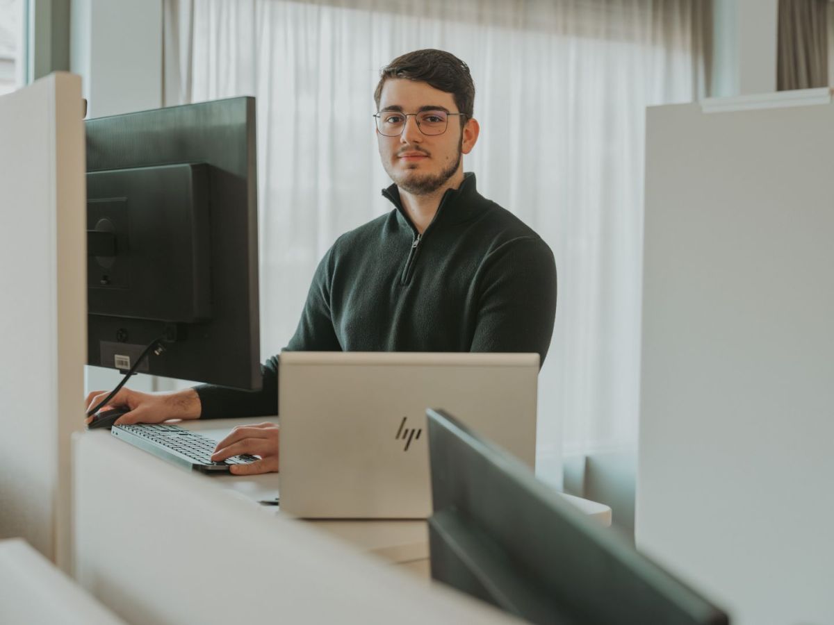 Junger Mann mit Brille sitzt an einem Schreibtisch und arbeitet am Computer; vor ihm stehen ein Monitor und ein Laptop in einem hellen Büro.