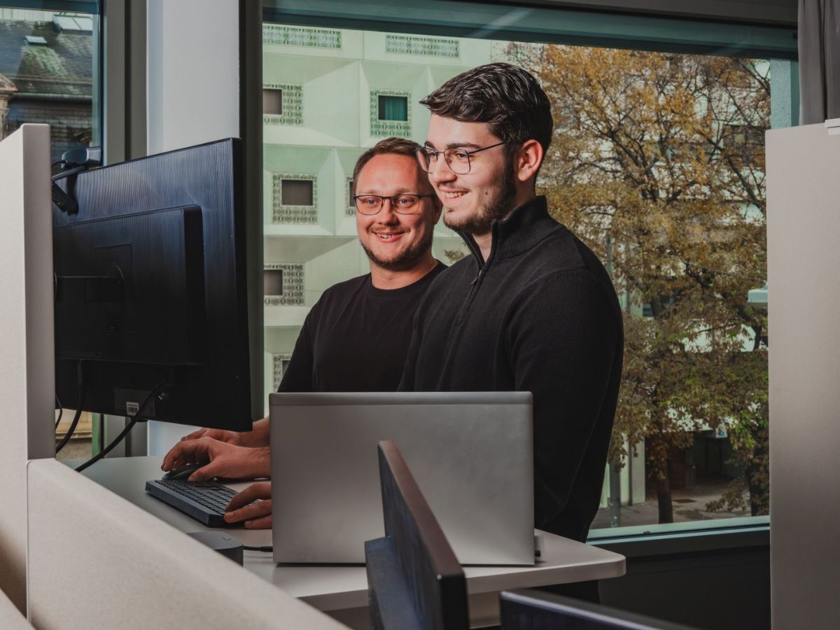 Zwei Männer stehen nebeneinander an einem Arbeitsplatz und arbeiten gemeinsam an einem Computer; beide schauen auf den Bildschirm, im Hintergrund ist ein Fenster mit Blick nach draussen.
