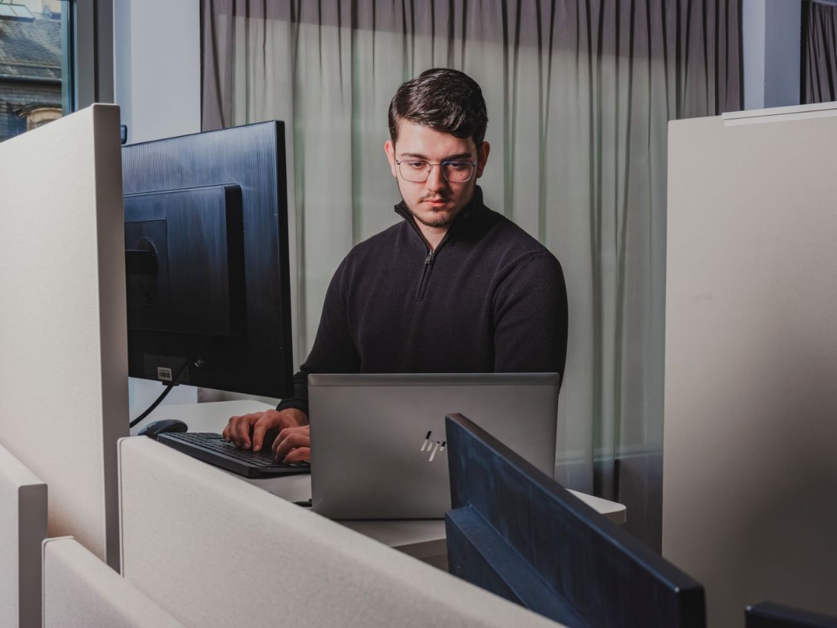 unger Mann mit Brille arbeitet konzentriert an einem Computerarbeitsplatz mit Monitor und Laptop in einem ruhigen Büro.