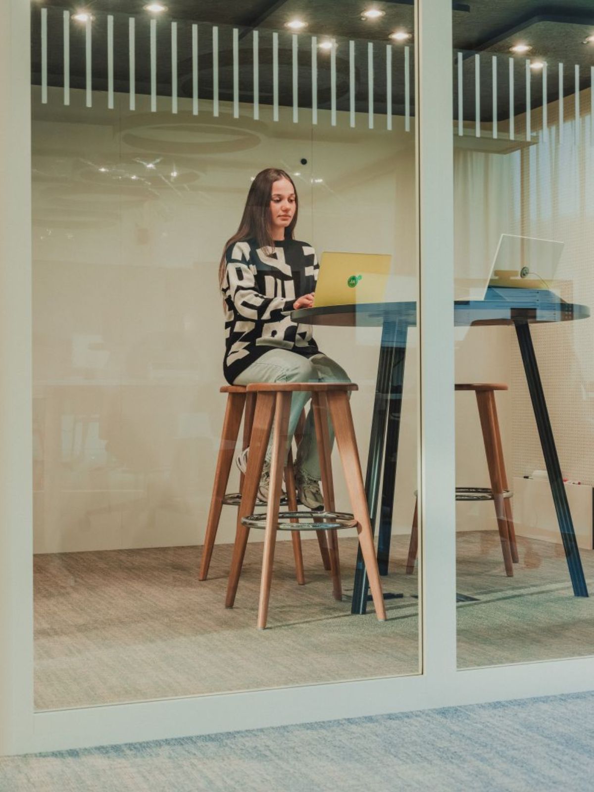 Junge Frau sitzt an einem hohen Tisch in einem gläsernen Besprechungsraum und arbeitet konzentriert an einem Laptop.
