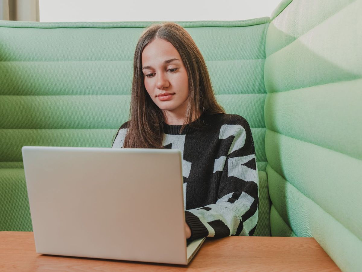 Junge Frau sitzt in einer grünen Sitznische und arbeitet konzentriert an einem Laptop an einem Tisch.
