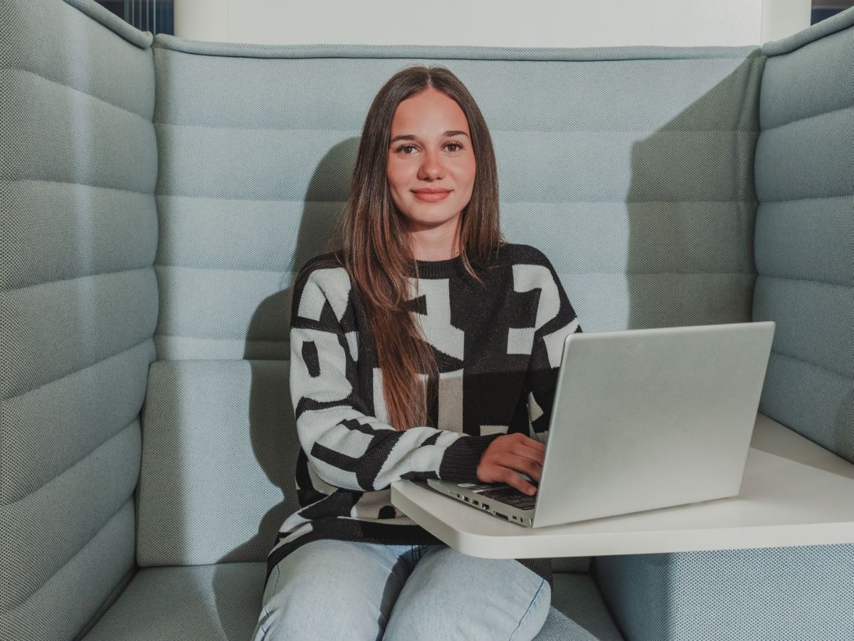 Junge Frau sitzt in einer Sitznische, arbeitet an einem Laptop und blickt lächelnd in die Kamera.