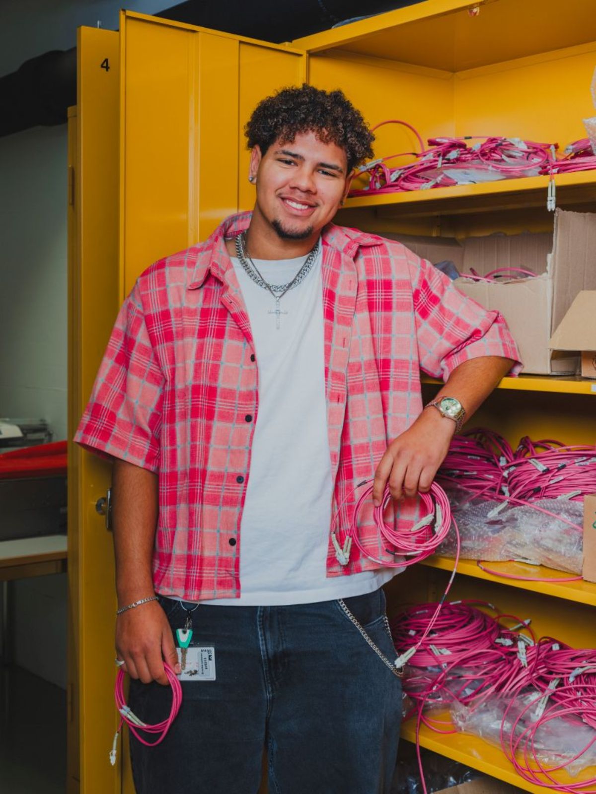 Junger Mann mit lockigem Haar steht lächelnd vor einem gelben Lagerschrank und hält pinke Kabel in der Hand; im Hintergrund sind weitere Kabel ordentlich gelagert.