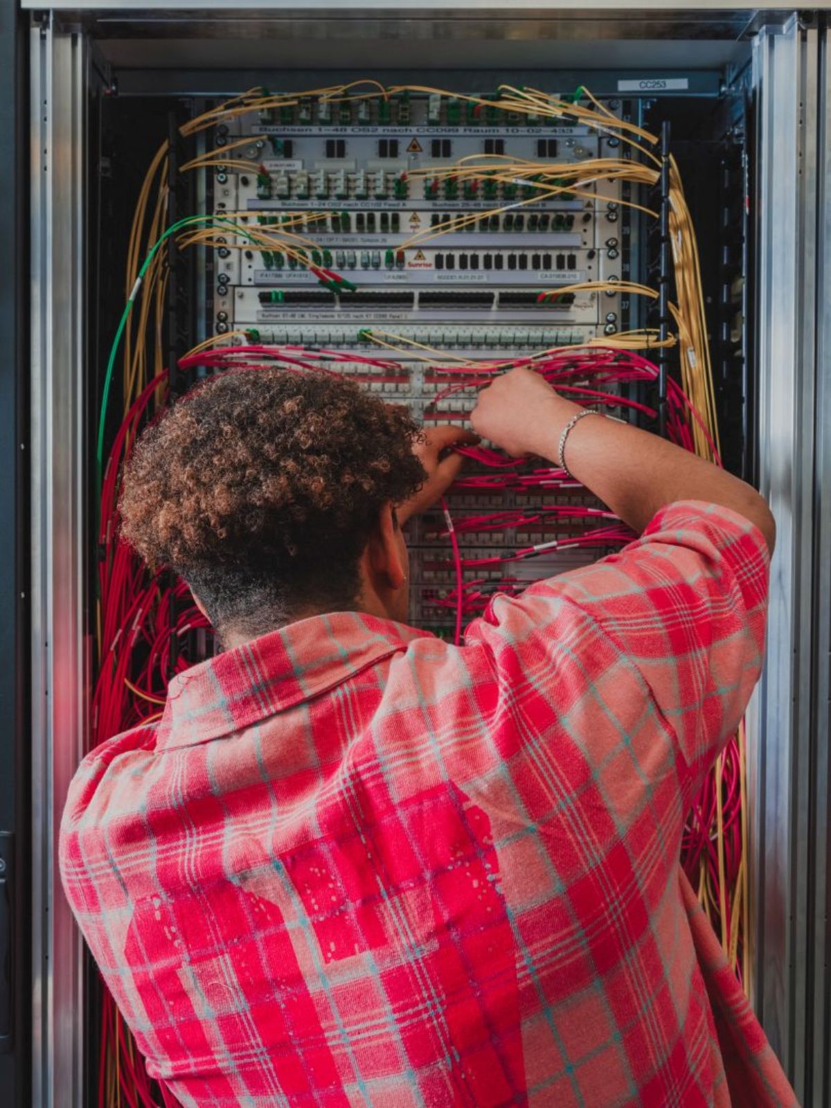 Junger Mann arbeitet an einem Netzwerkschrank und verbindet rote und gelbe Kabel; zu sehen ist die Rückansicht während er konzentriert die Verkabelung ordnet.
