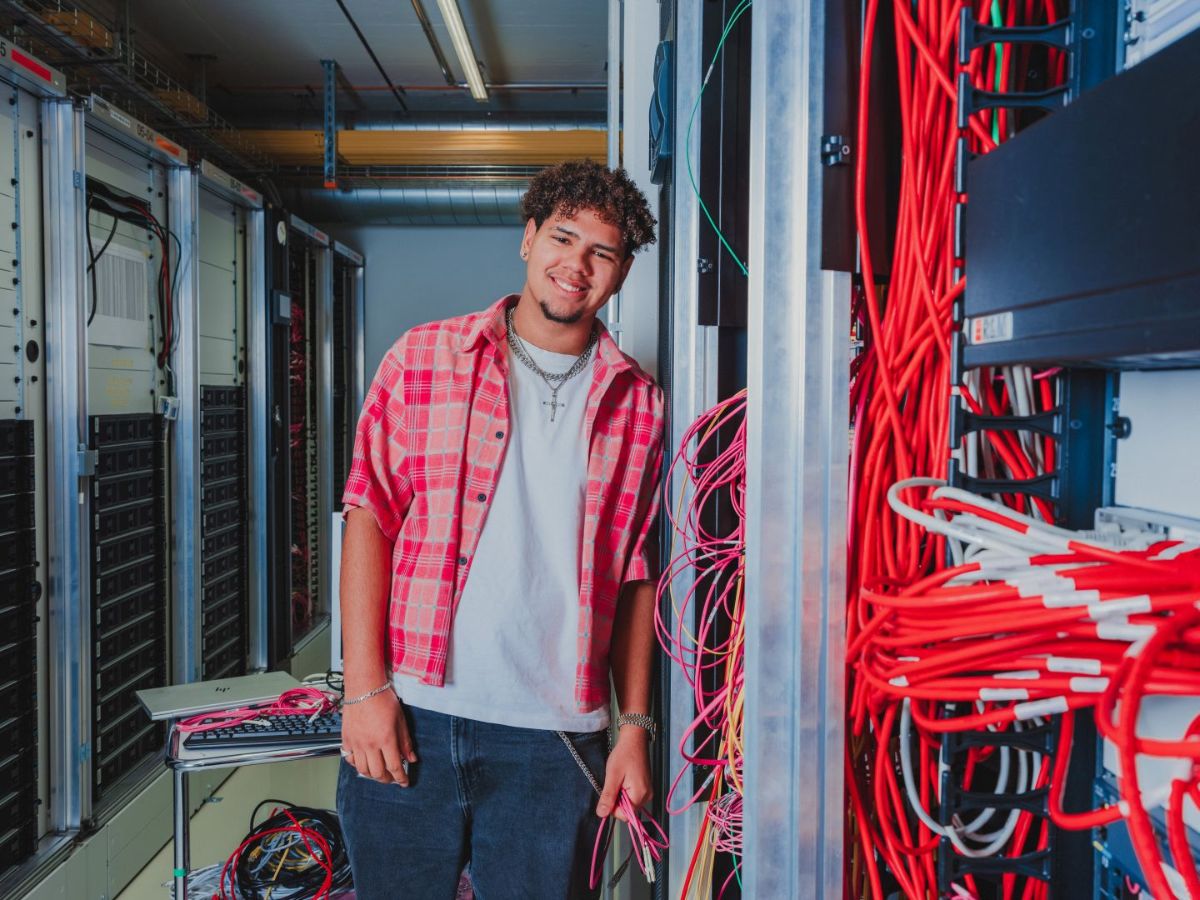 Junger Mann steht lächelnd in einem Serverraum zwischen Netzwerkschränken und hält pinke Kabel in der Hand; im Hintergrund sind zahlreiche rote und schwarze Kabel installiert.