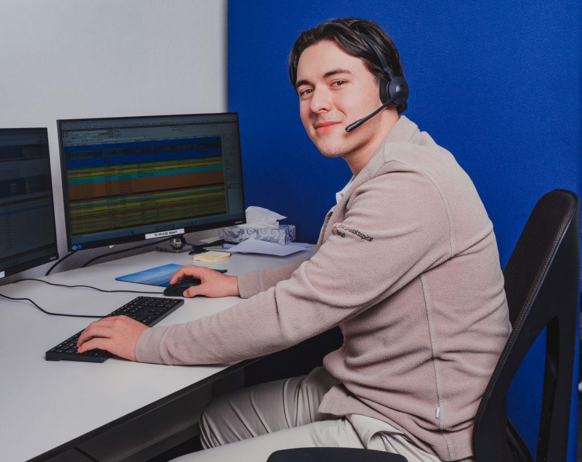 Junger Mann mit Headset sitzt an einem Büroarbeitsplatz vor zwei Monitoren und arbeitet am Computer; er blickt lächelnd zur Kamera.