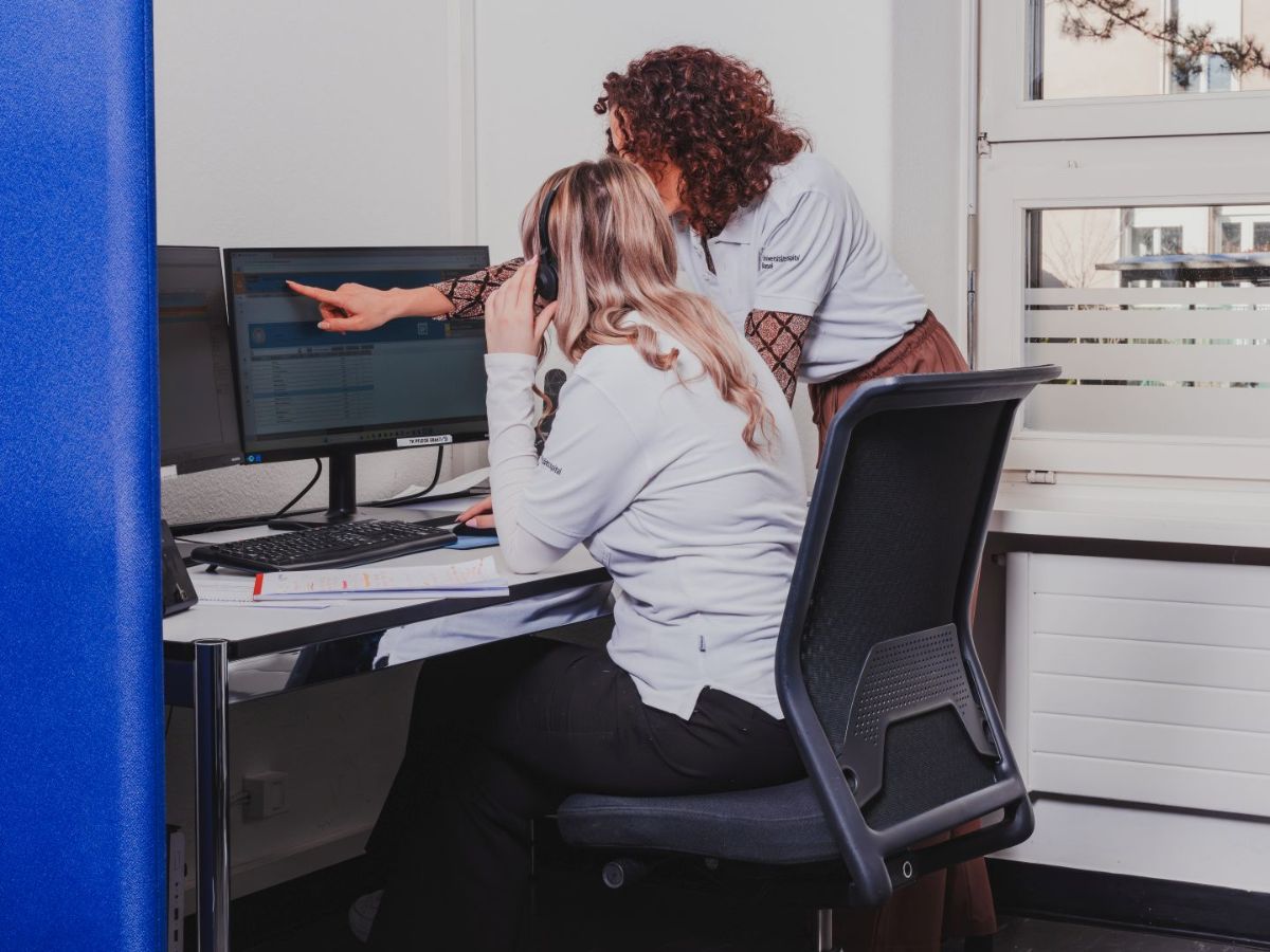 Zwei Frauen arbeiten gemeinsam an einem Computerarbeitsplatz; eine sitzt mit Headset vor dem Bildschirm, während die andere daneben steht und auf den Monitor zeigt.