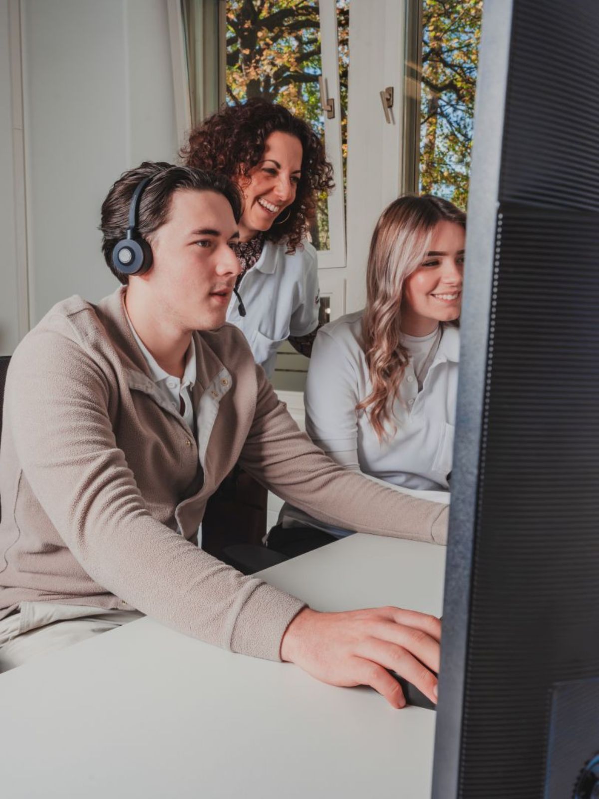 Drei junge Personen arbeiten gemeinsam am Computer: Eine Person mit Headset sitzt am Bildschirm und bedient die Maus, während zwei Kolleginnen danebenstehen, lächeln und unterstützend mitarbeiten; im Hintergrund fällt Tageslicht durch ein Fenster.