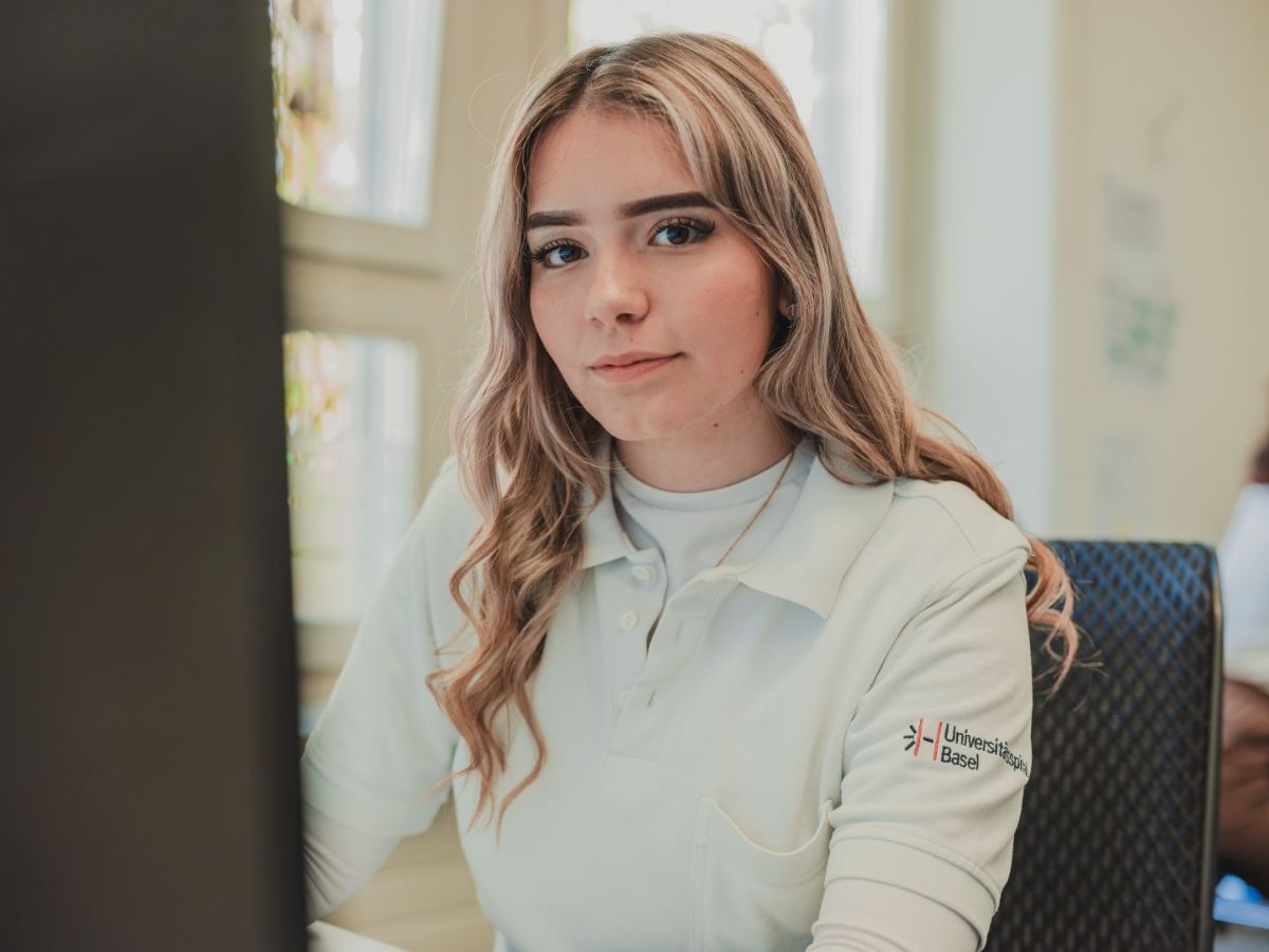 Junge Frau sitzt an einem Computerarbeitsplatz und blickt aufmerksam in die Kamera; sie trägt ein helles Oberteil mit Logo.