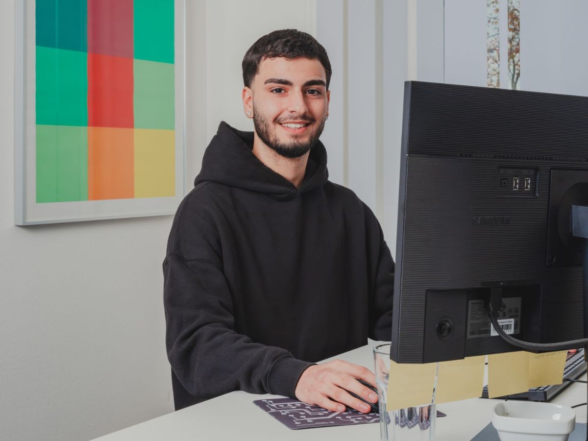 Junger Mann sitzt an einem Computerarbeitsplatz und blickt lächelnd in die Kamera; vor ihm stehen Monitor und Maus auf einem Schreibtisch.