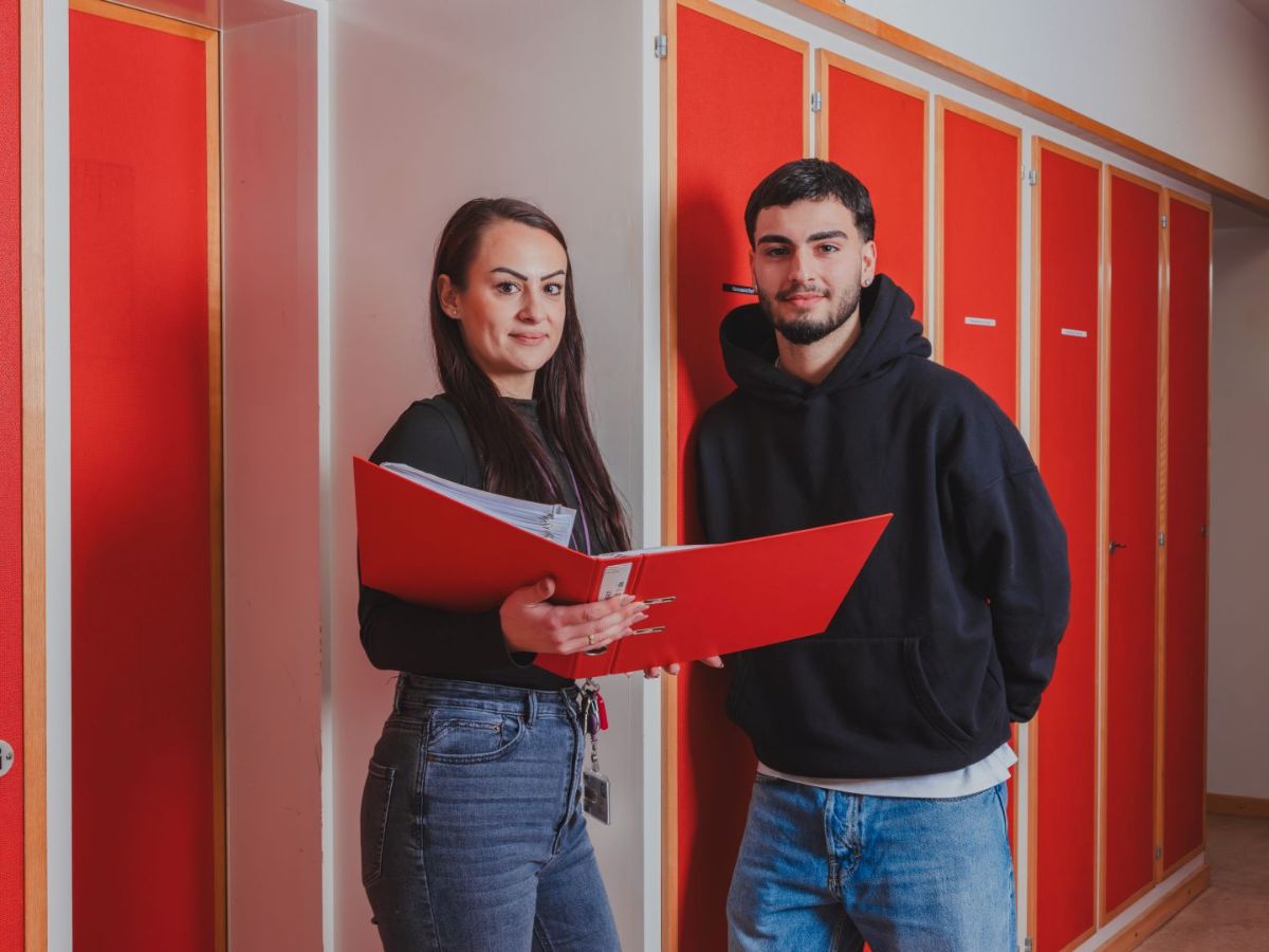 Zwei Personen stehen in einem Flur vor roten Schranktüren; eine hält einen geöffneten Ordner, während beide in die Kamera blicken.